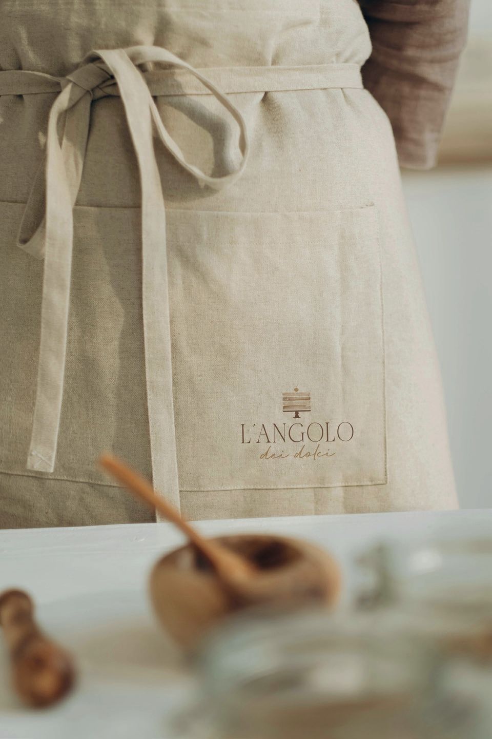 Grembiule di lino beige annodato in vita, con tasca con logo; cucchiaio di legno e ciotola in primo piano.