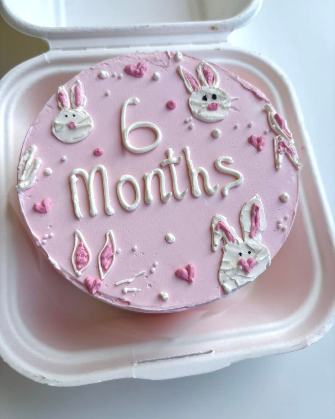 Torta rosa con decorazioni "6 mesi" e coniglietto in un contenitore bianco.