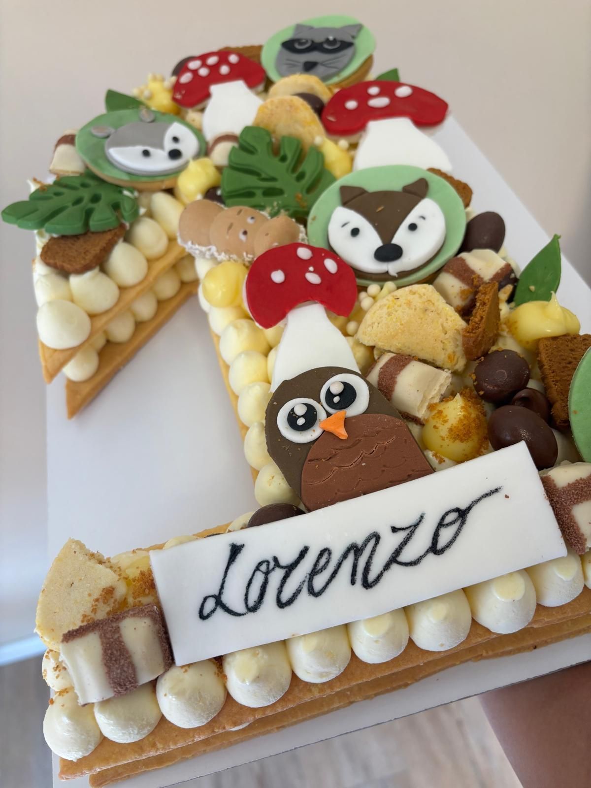 Dolce a forma di lettera, decorato con bacche, cioccolato e fiori su una superficie dorata.