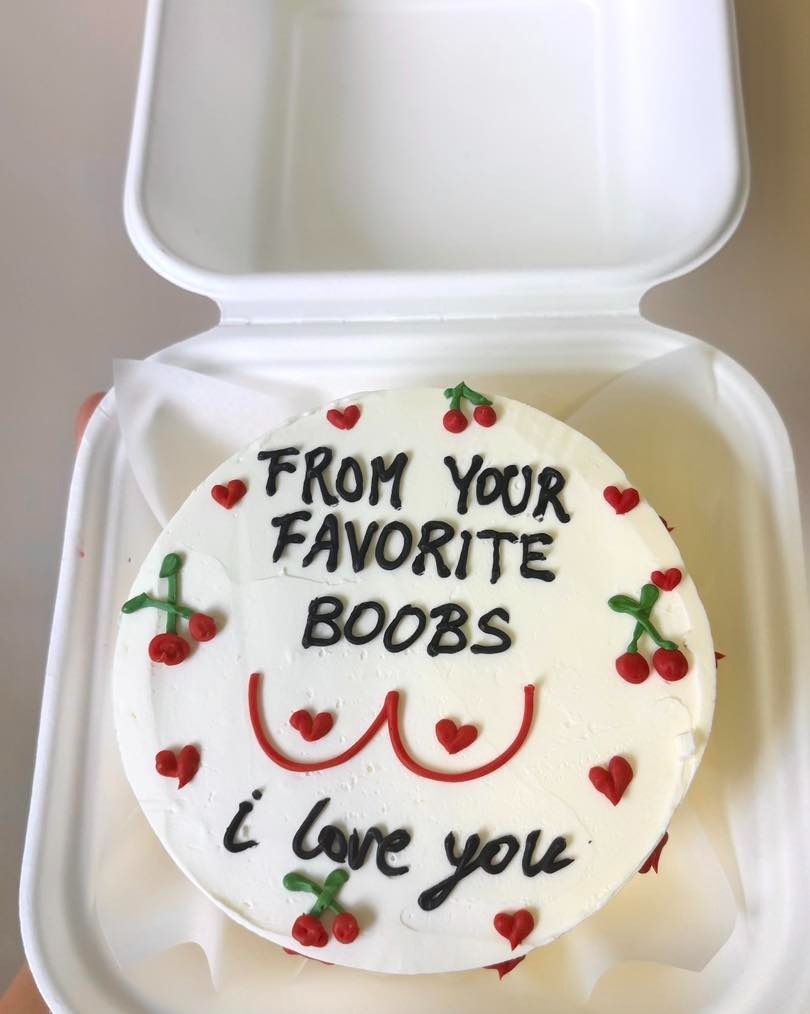 Torta bianca con la scritta "Dal tuo seno preferito, ti amo", decorata con ciliegie e cuori, in un contenitore da asporto.