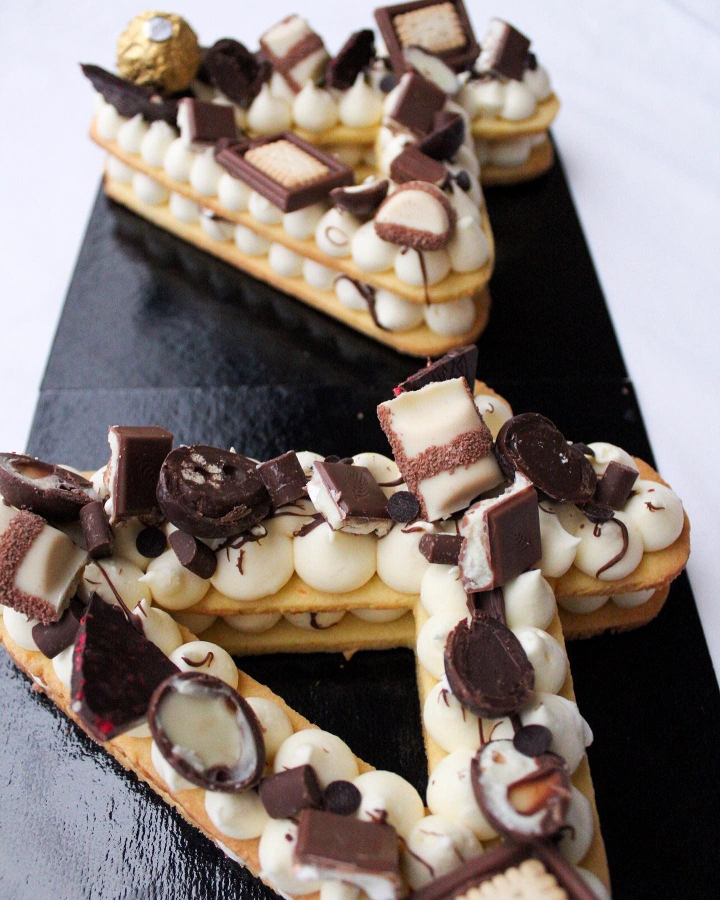 Dessert ricoperto di cioccolato a forma di lettera "V" su un vassoio nero, con glassa alla crema e cioccolatini.