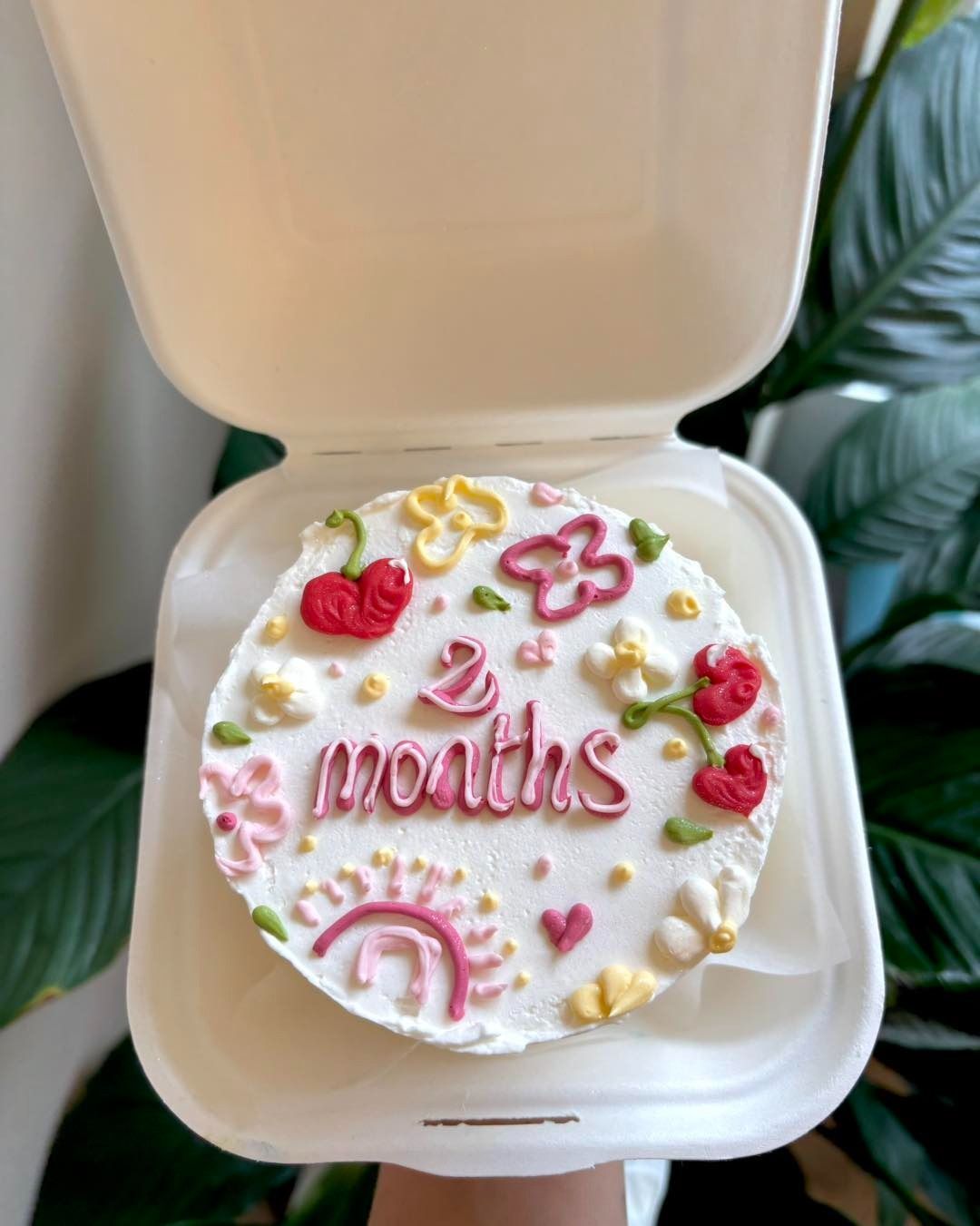 Piccola torta bianca decorata con la scritta "2 mesi" e fiori, in un contenitore da asporto.