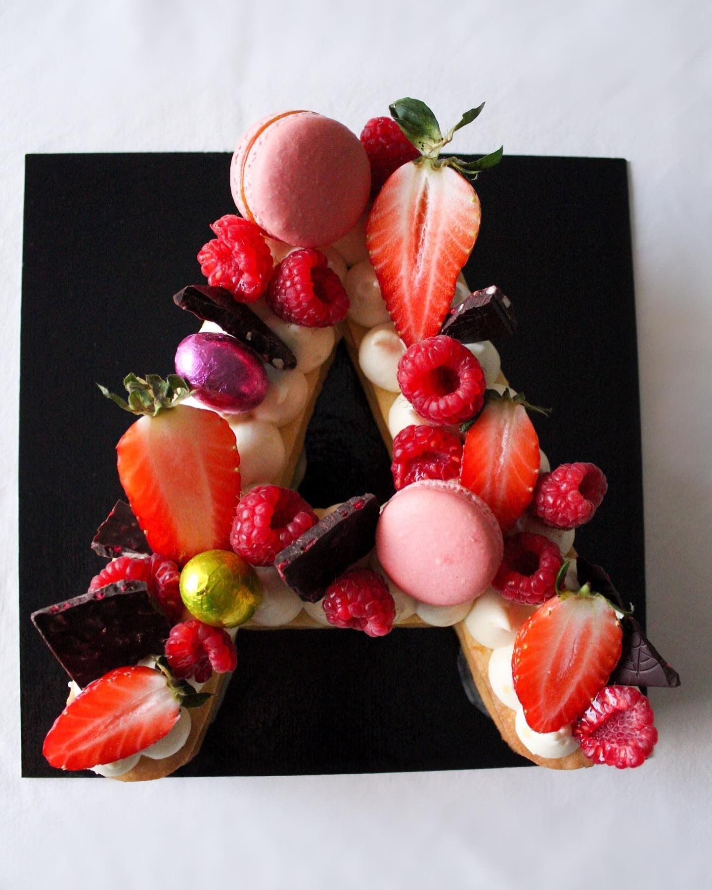 Torta a forma di lettera "A" decorata con frutti di bosco, macaron e cioccolato su un piatto quadrato nero.