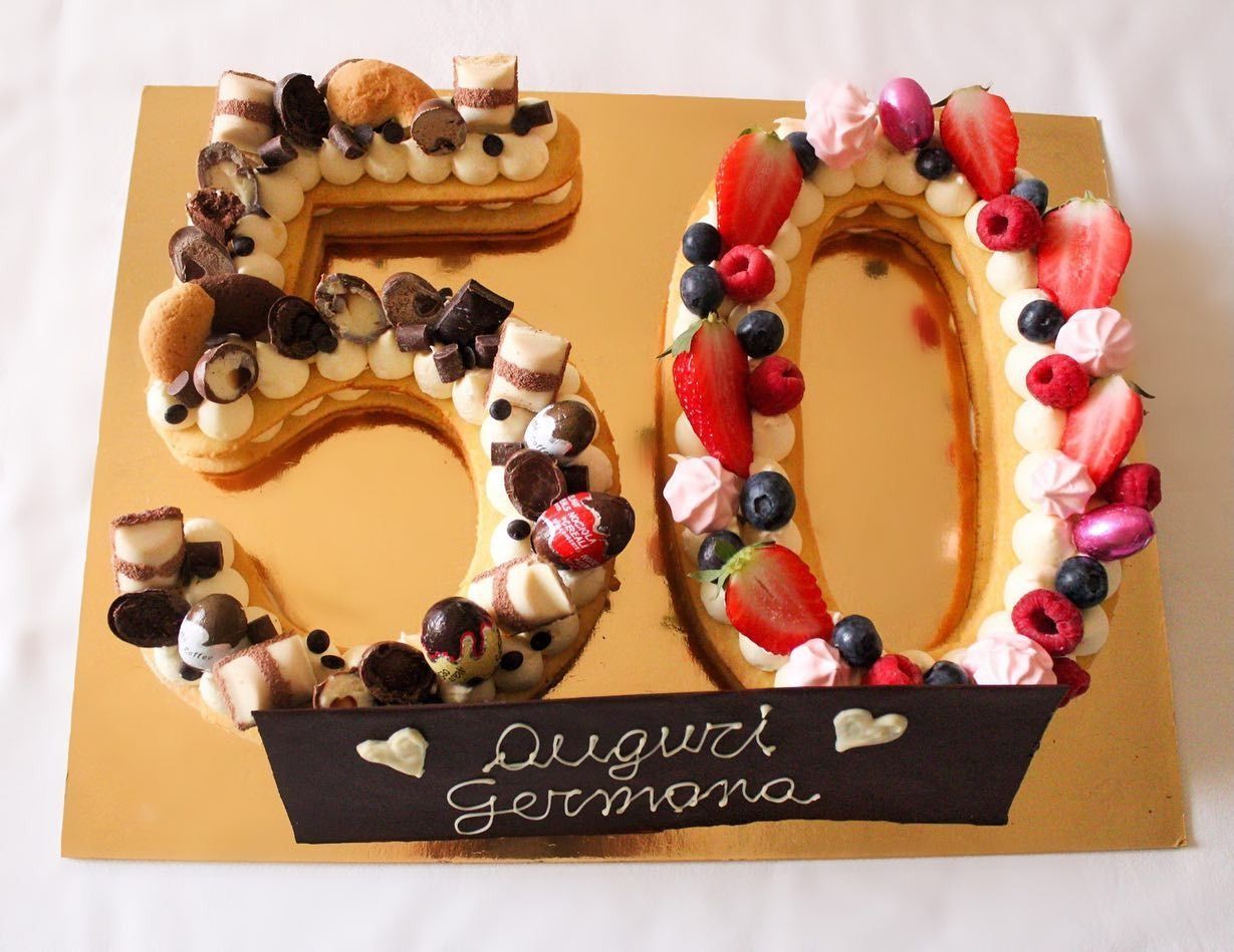 Torta a forma di numero 50, decorata con panna, frutta e cioccolato. Sopra c'è la scritta "Auguri Germana".