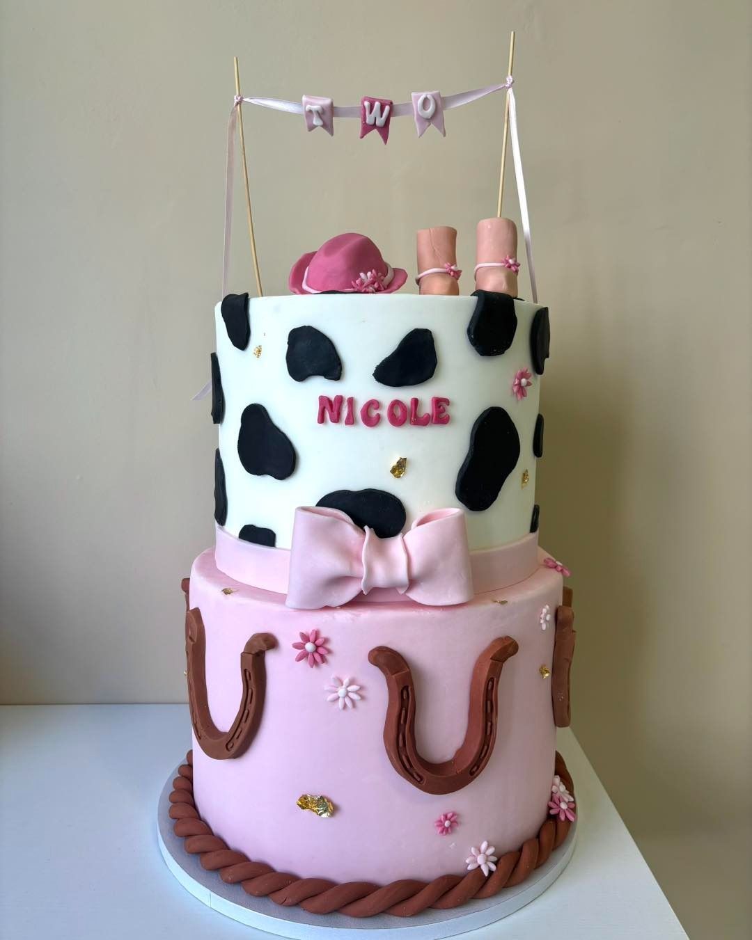 Torta a due piani a tema cowgirl con motivi rosa e in pelle di mucca, decorata con un cappello, stivali e uno striscione.