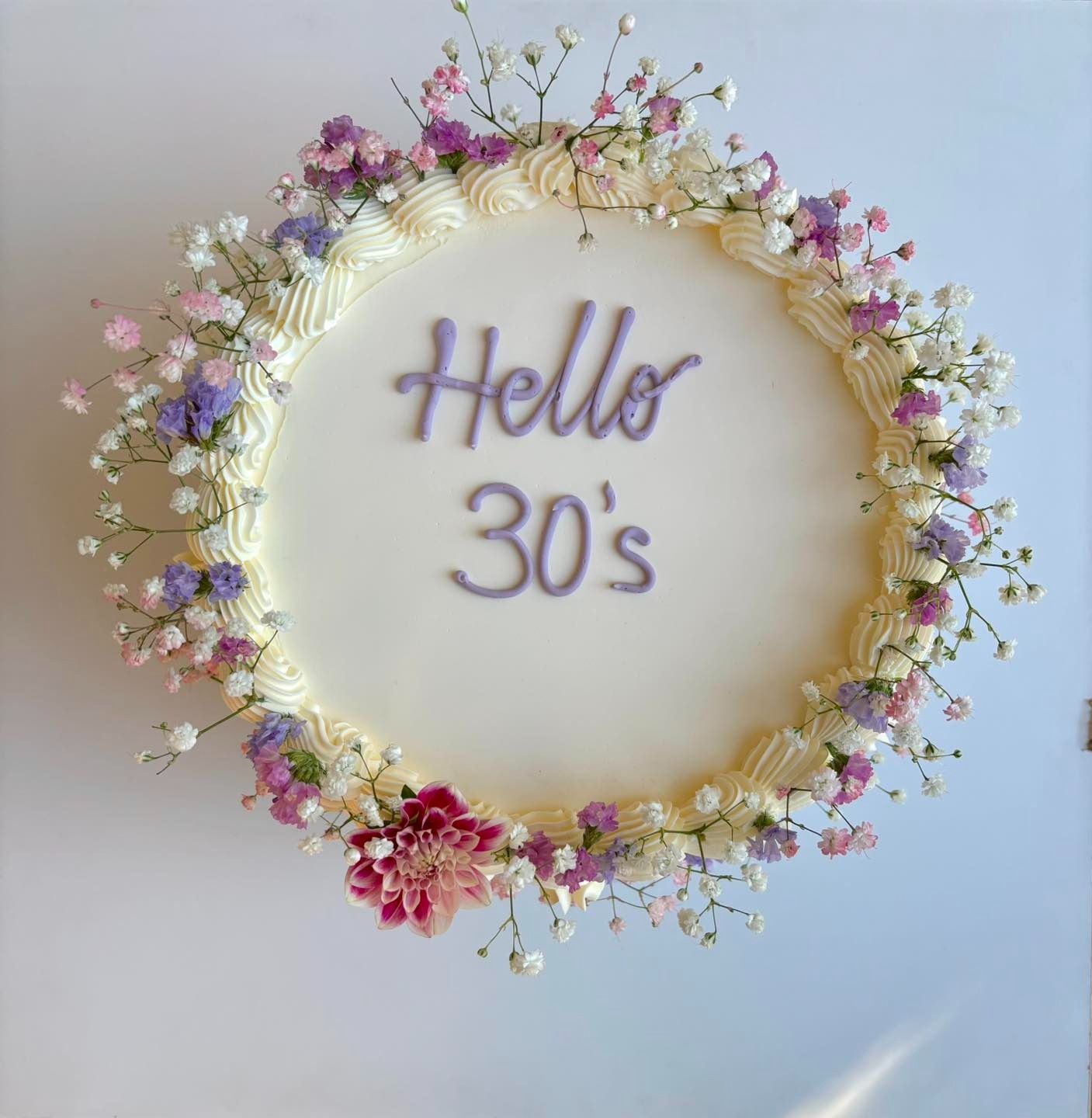 Torta decorata con fiori, scritta "Hello 30's" con glassa viola su una torta glassata bianca.