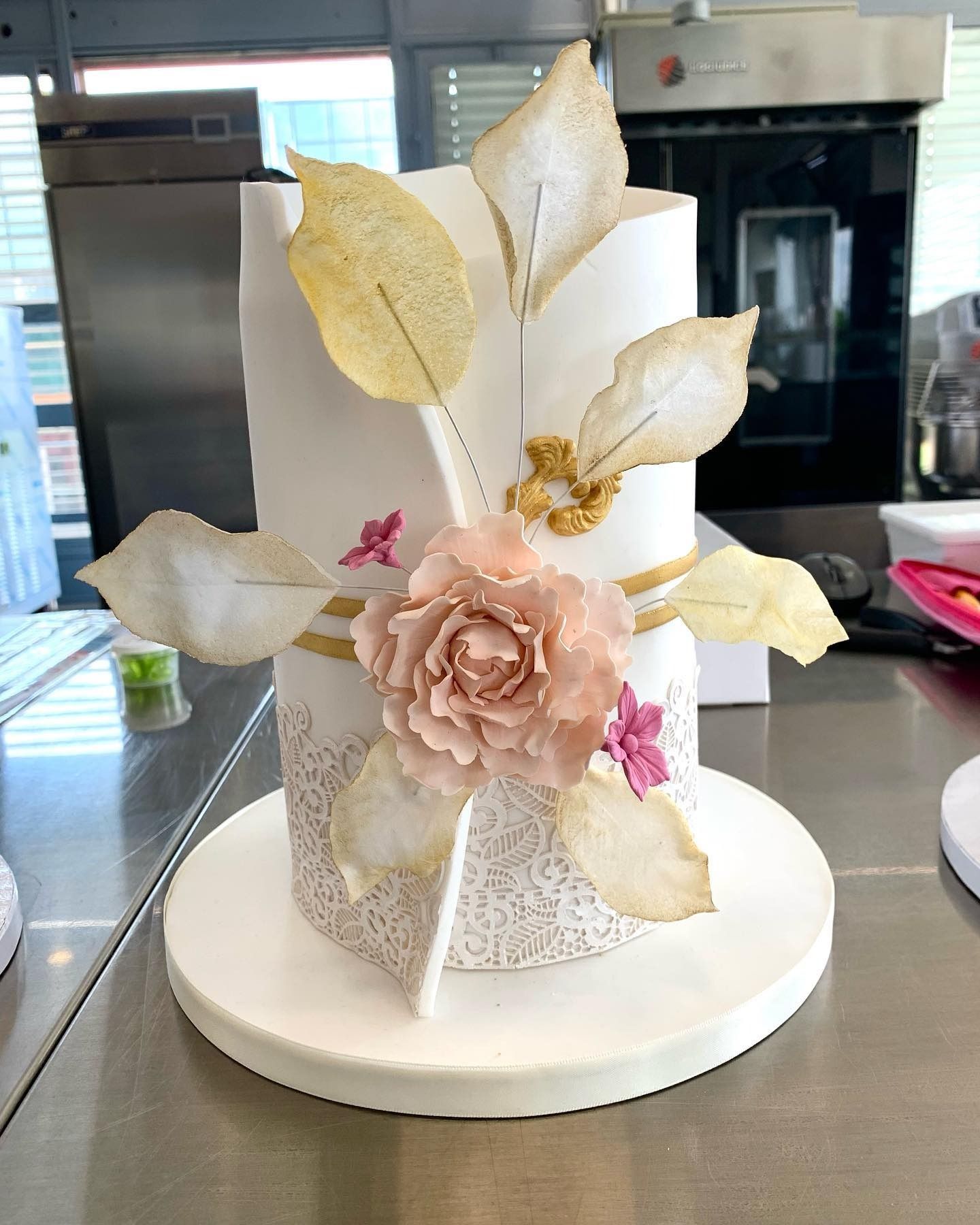 Torta bianca a due piani con dettagli dorati, fiori di zucchero e foglie. Posizionata su un'alzata bianca.