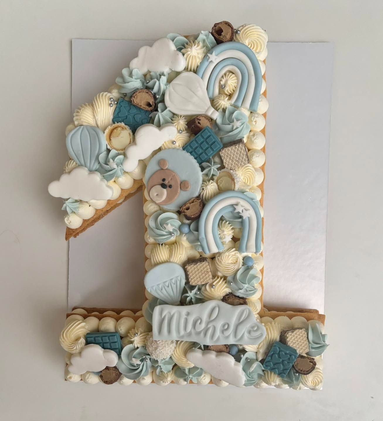 Torta numero "1" decorata con glassa blu e bianca, caramelle e decorazioni a forma di orsacchiotto.