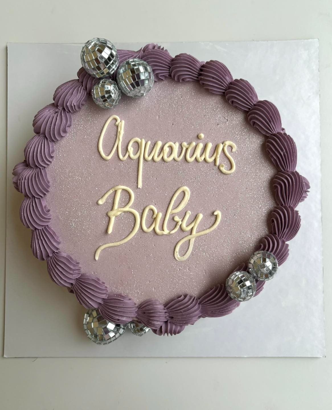 Torta alla lavanda con la scritta "Aquarius Baby" in bianco e dettagli a forma di palla da discoteca.