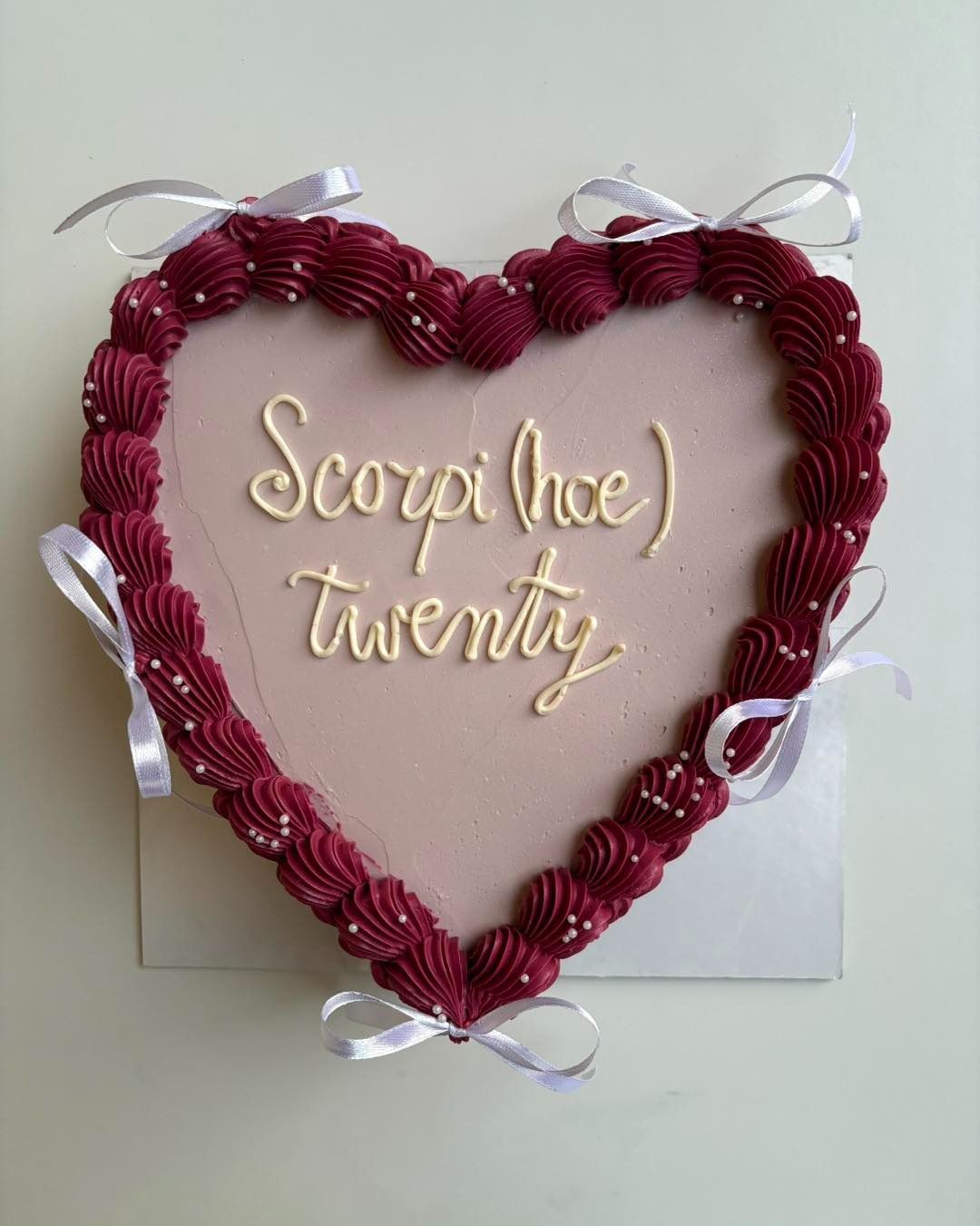 Torta a forma di cuore con bordo di glassa bordeaux e scritta "Scorpi(hoe) twenty" in crema. Fiocchi di nastro bianco come decorazione.