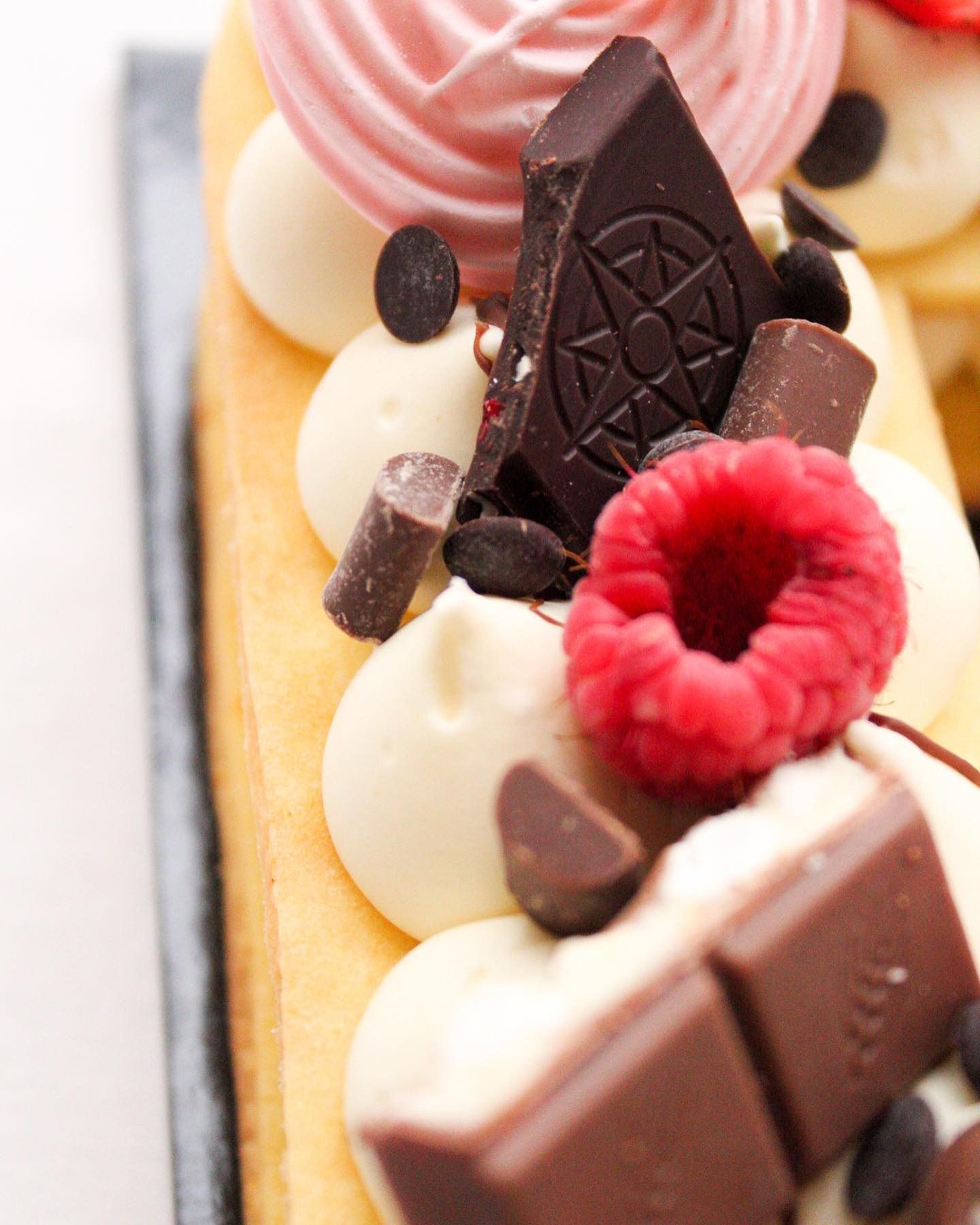 Primo piano di un dessert: torta con glassa bianca, gocce di cioccolato, lamponi e meringa rosa.