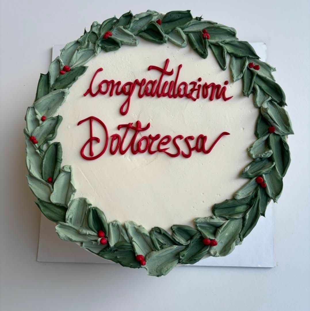 Torta con la scritta "Congratulazioni Dottoressa" in rosso, circondata da una ghirlanda di foglie verdi, su glassa bianca.