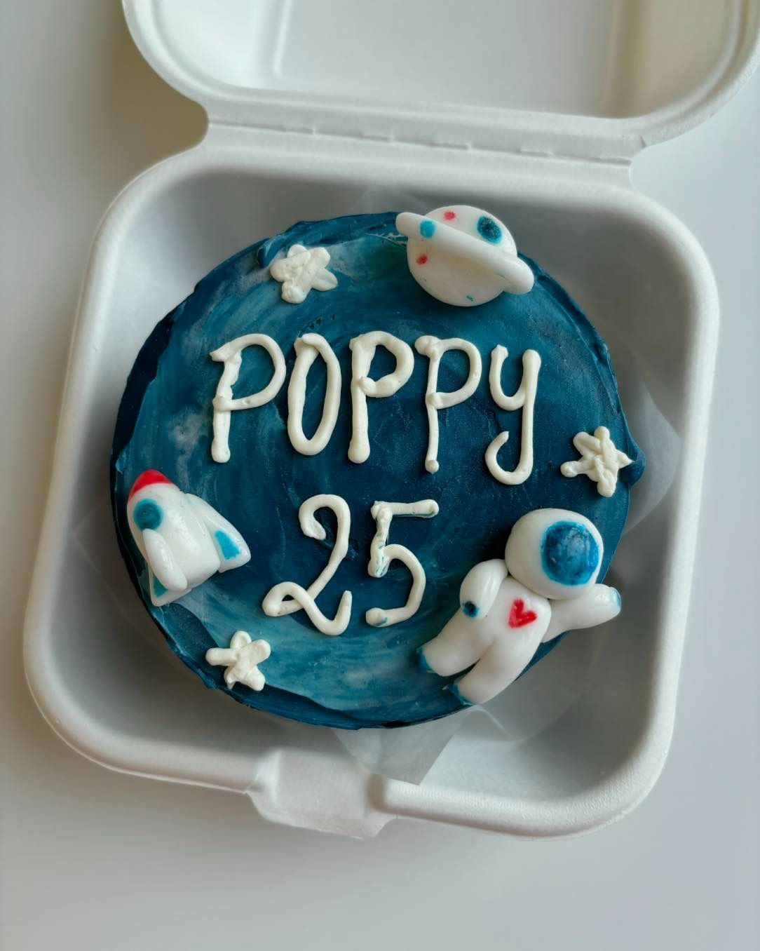 Torta in un contenitore, decorata con un motivo a spirale di glassa blu, testo bianco "POPPY 25" e pasta di zucchero a tema spaziale.