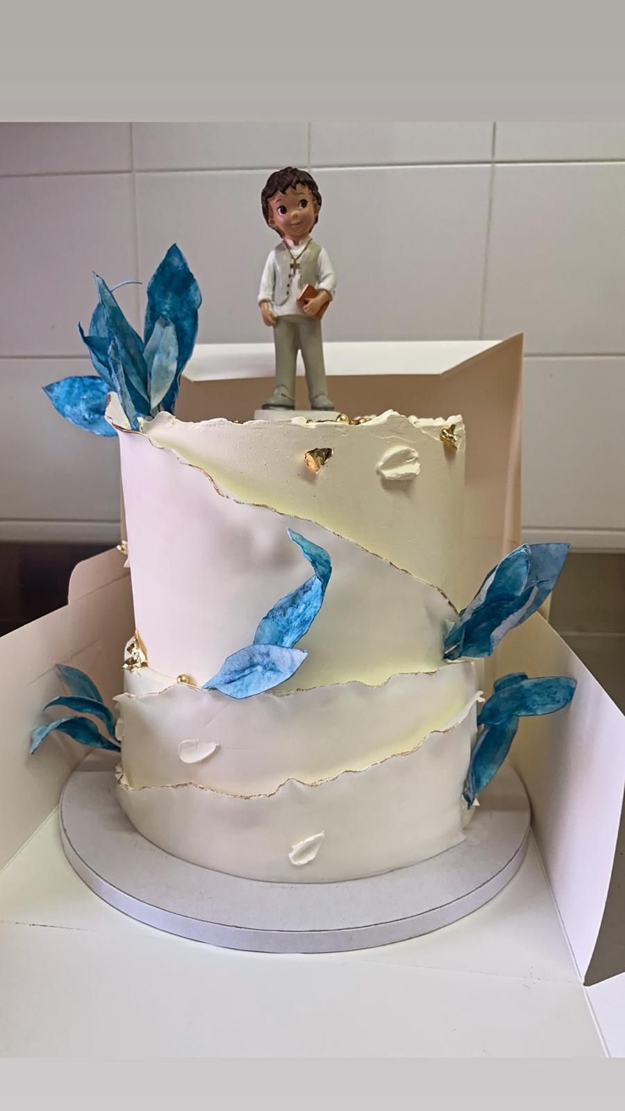 Torta bianca a due piani con dettagli dorati, fiori di zucchero e foglie. Posizionata su un'alzata bianca.