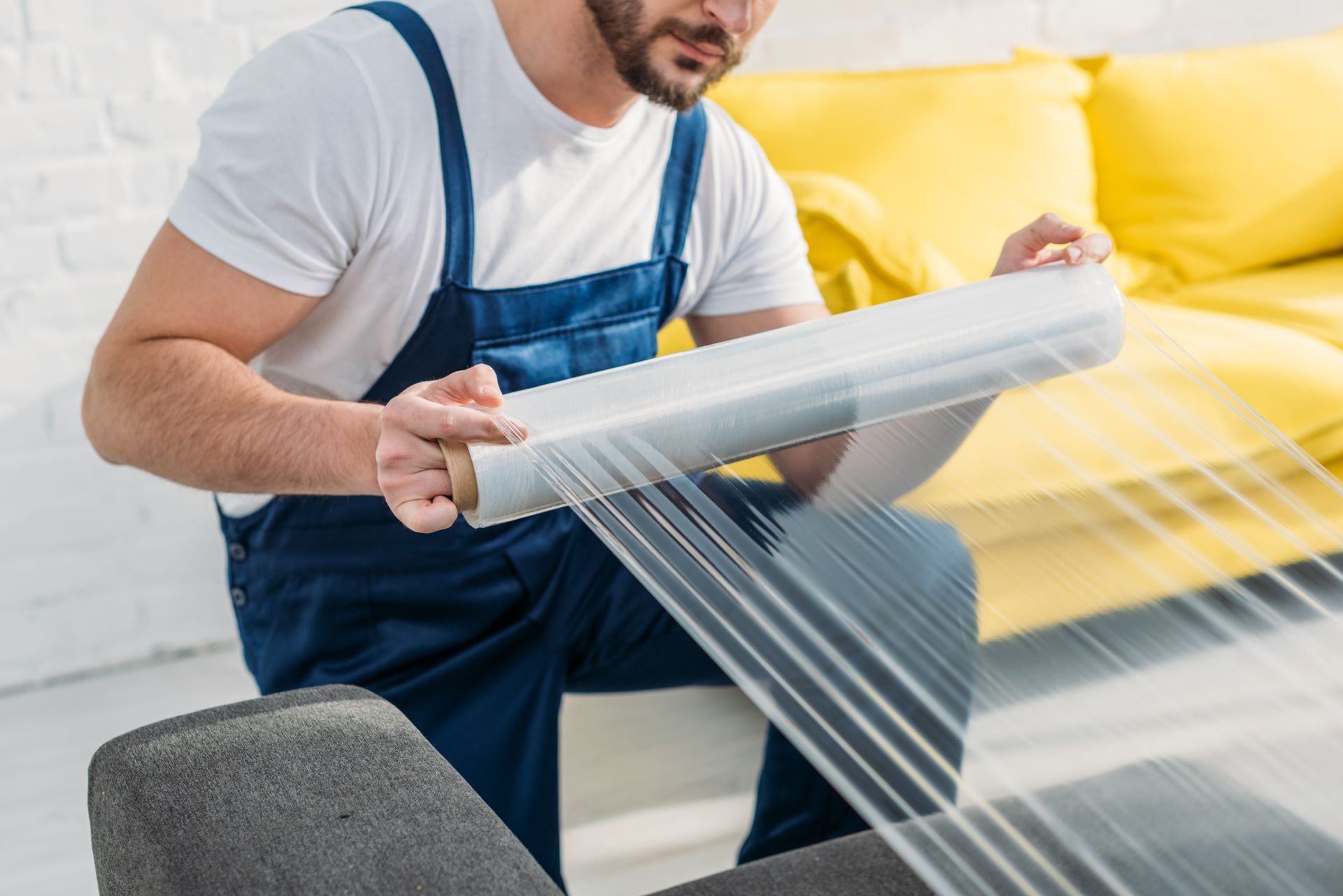 Man in blue overalls wrapping a chair with clear plastic wrap.