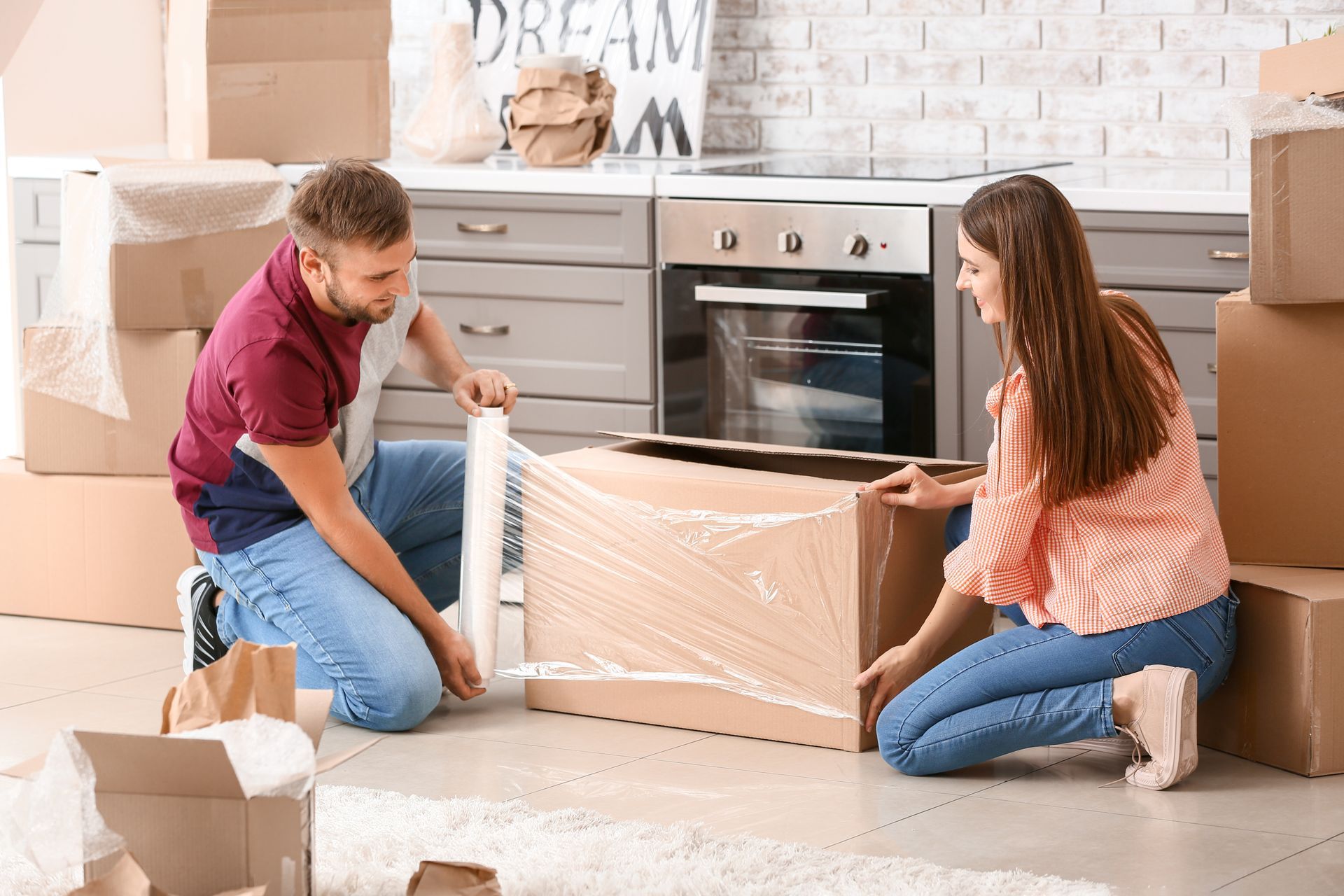 A couple packing cardboard boxes in a kitchen. They are using plastic wrap.