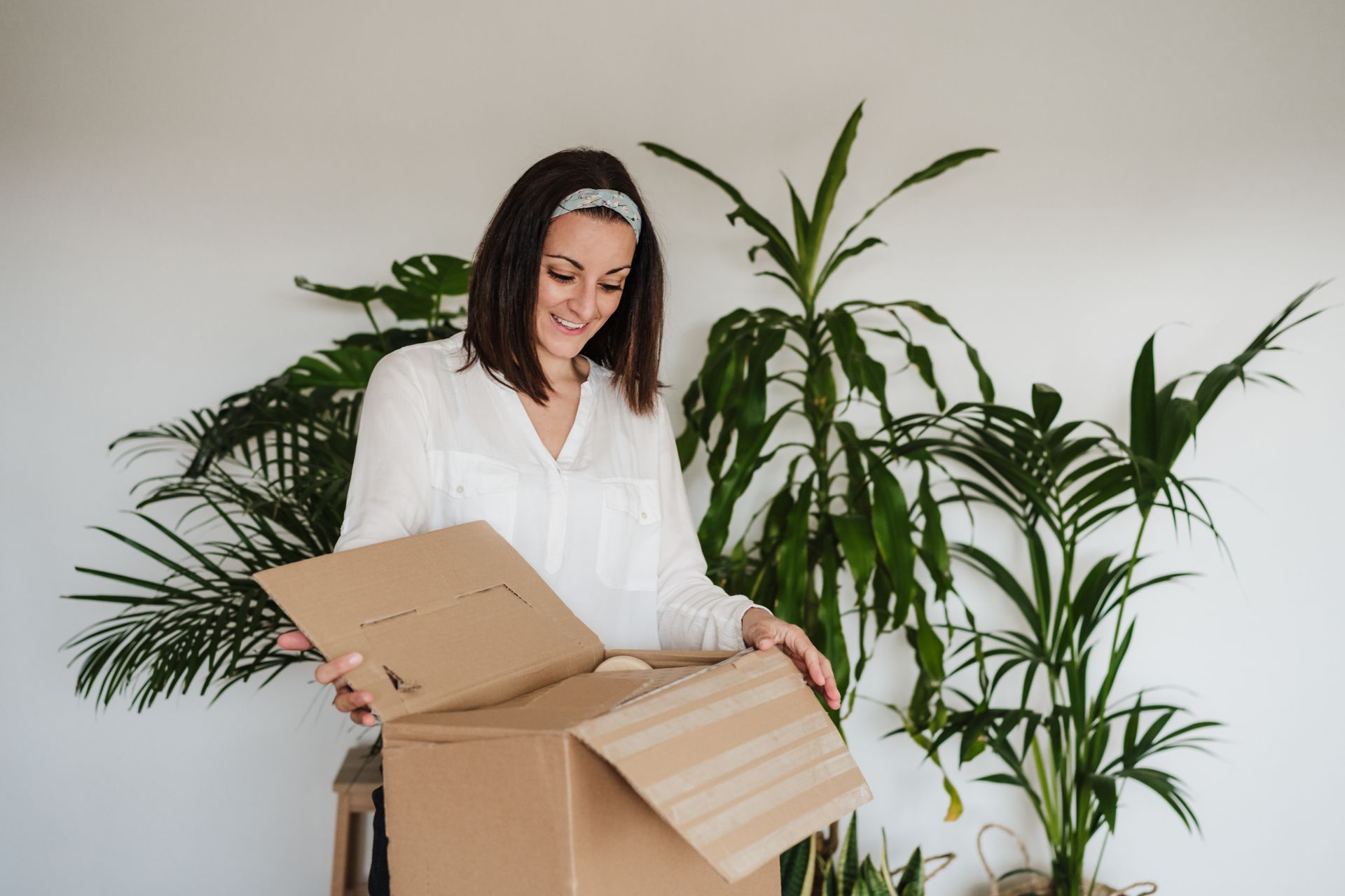 Woman opens a cardboard box with plants in the background; smiling in a bright room.