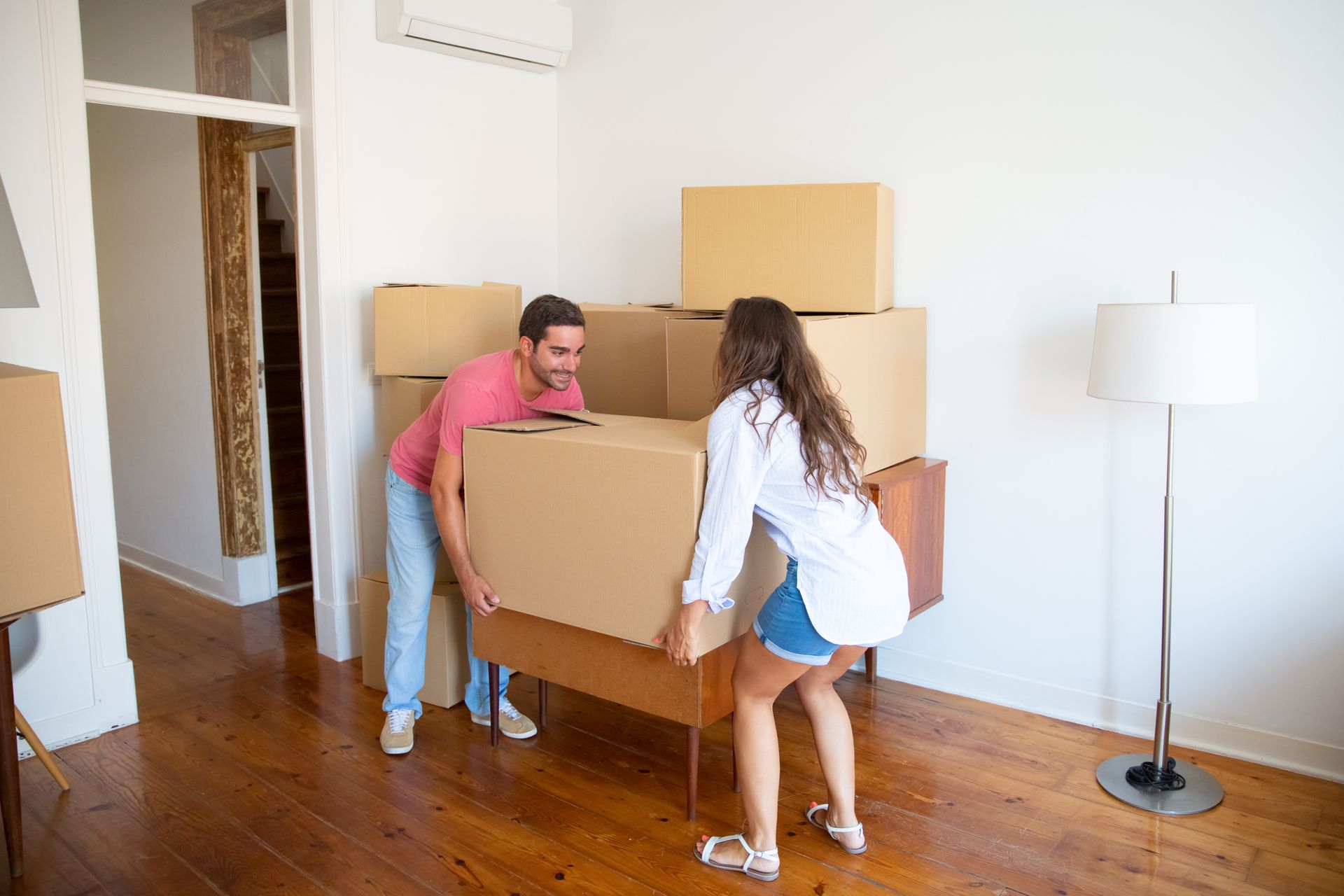 Couple lifting a large cardboard box onto a wooden dresser in a room with boxes and a lamp.