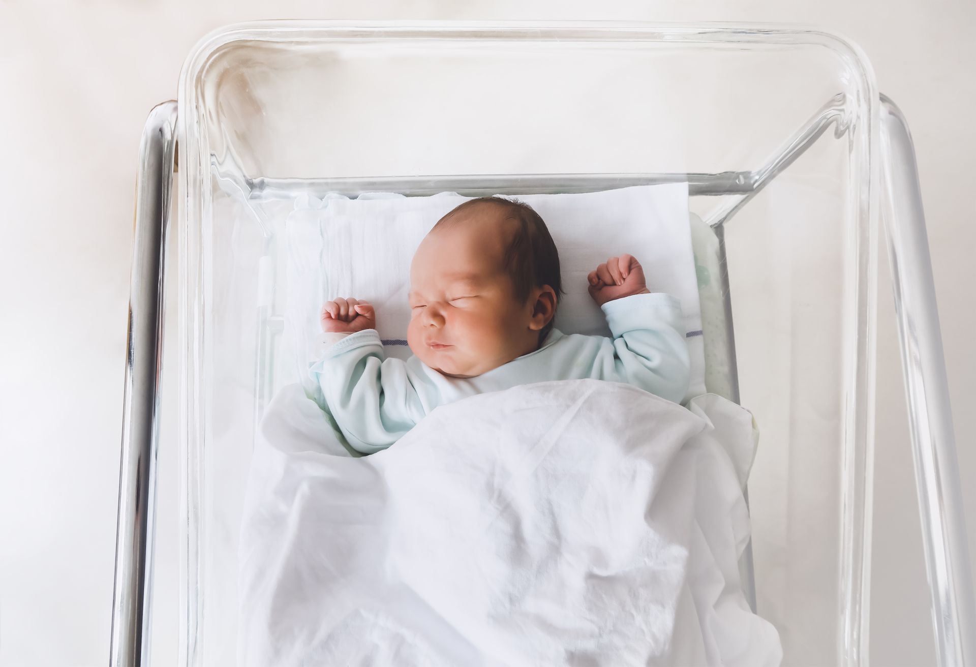 A newborn baby sleeping on its back in a clear bassinet.
