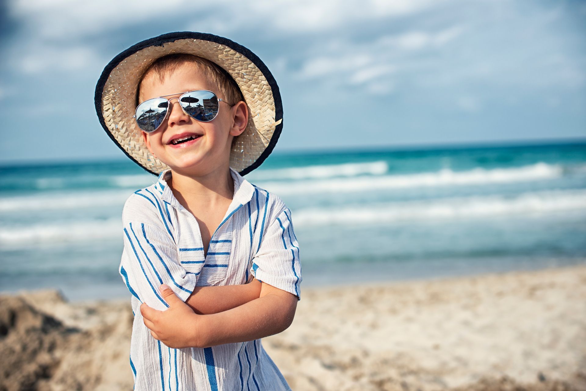 A young boy smiling, standing on a beach wearing a sun hat and striped shirt.