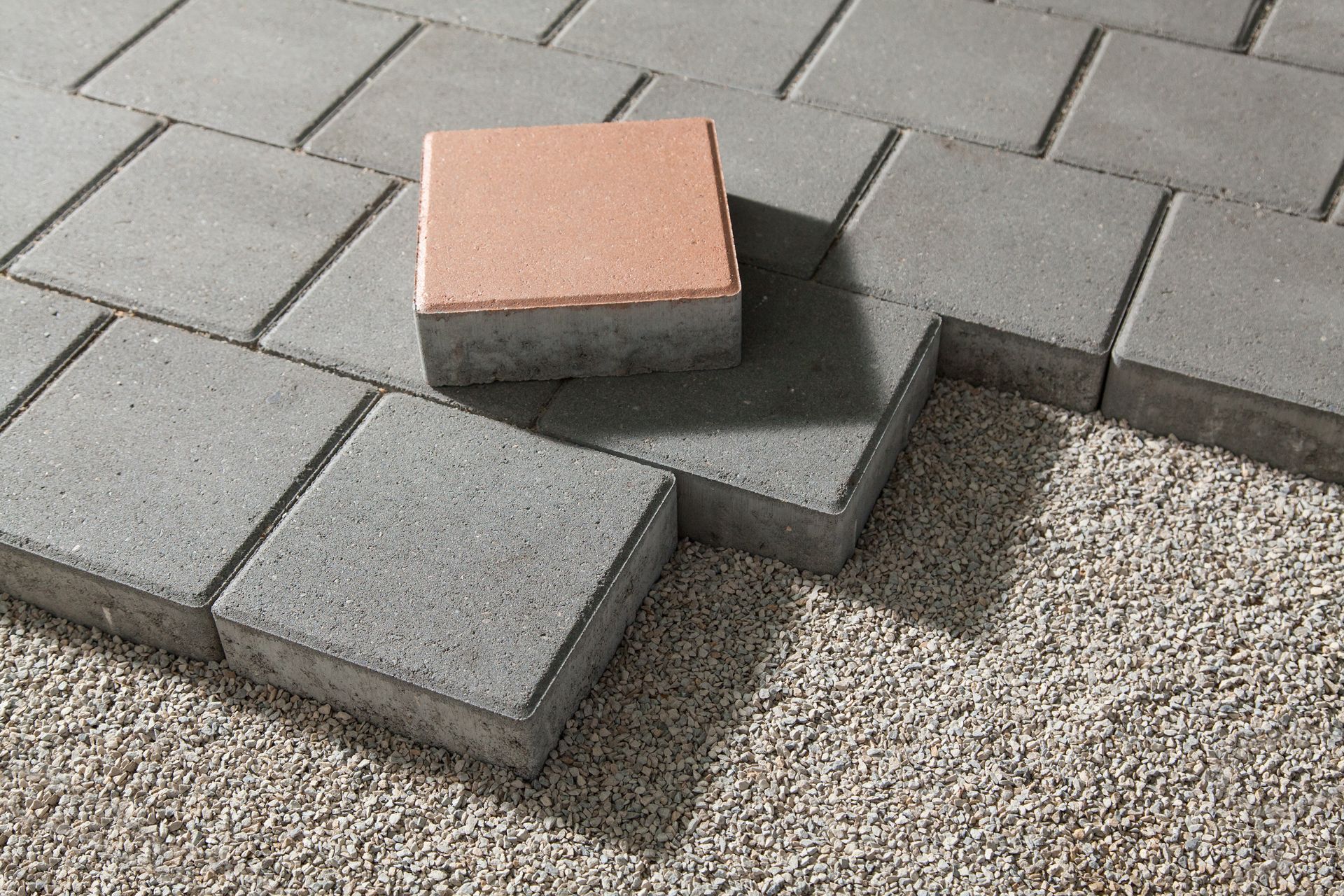 A light-colored square paver sits atop a pattern of gray concrete pavers partially installed on a bed of gravel.