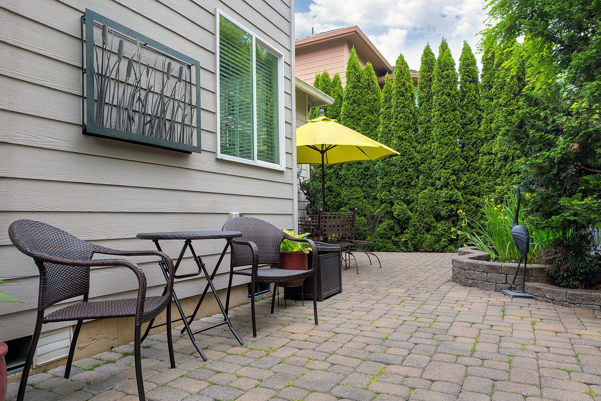 Patio with black metal table and chairs beside a house, yellow umbrella, and green hedge. Patio with black metal table and chairs beside a house, yellow umbrella, and green hedge.