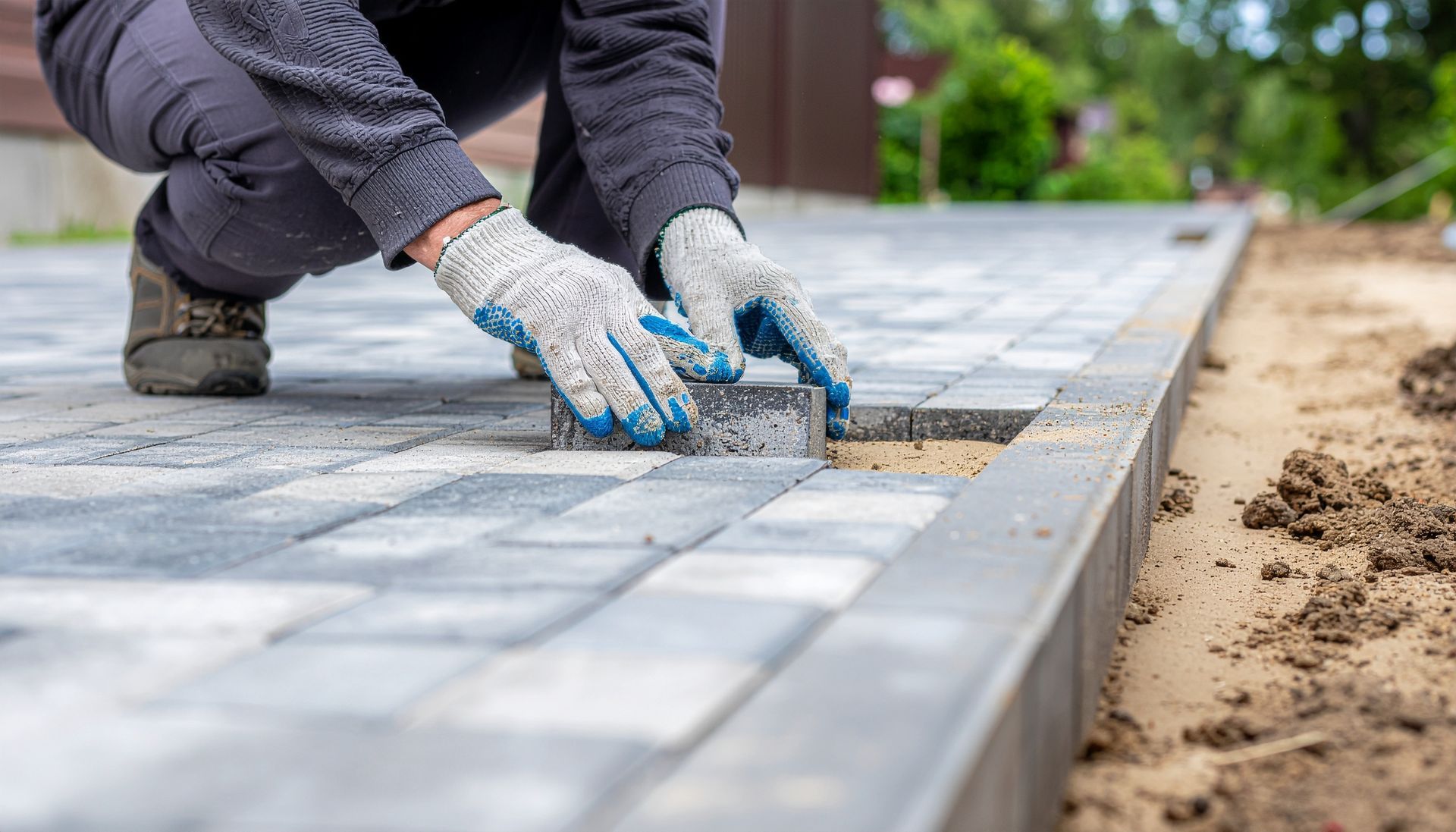 A person in work gloves laying gray paving stones into a sand base to complete a walkway.