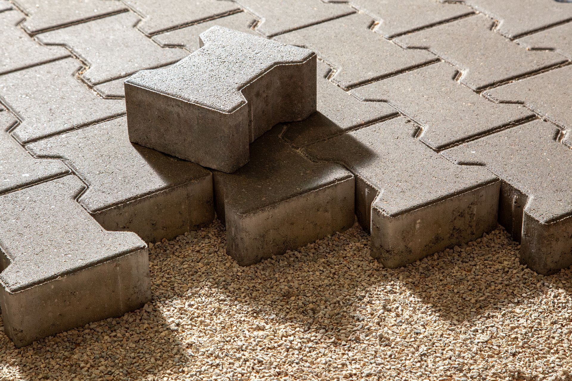 An I-shaped concrete paver sits on top of a partially laid gray interlocking pavement pattern over a gravel base.