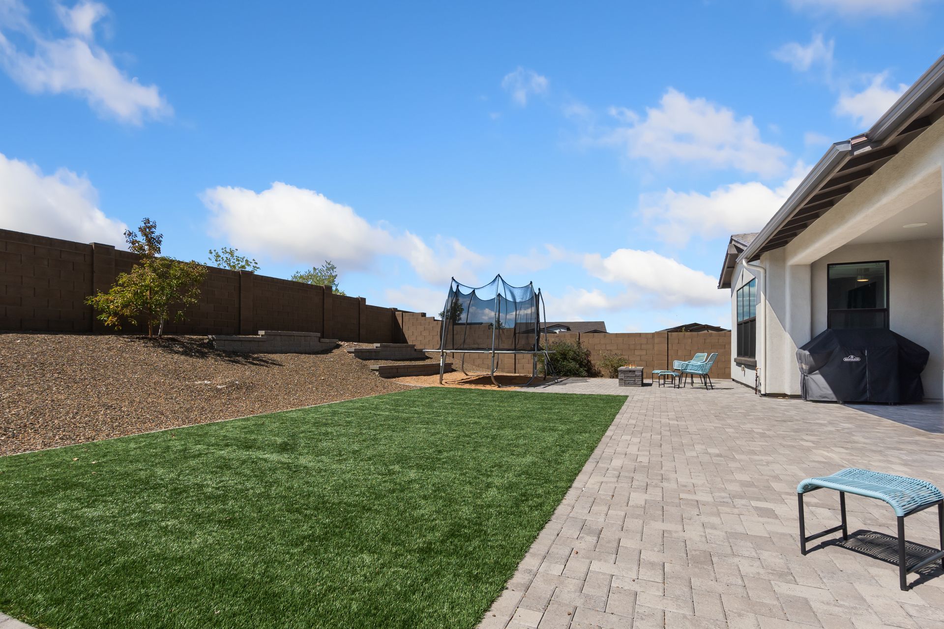Backyard with green lawn, paver patio, trampoline, and covered house patio under a blue sky Backyard with green lawn, paver patio, trampoline, and covered house patio under a blue sky