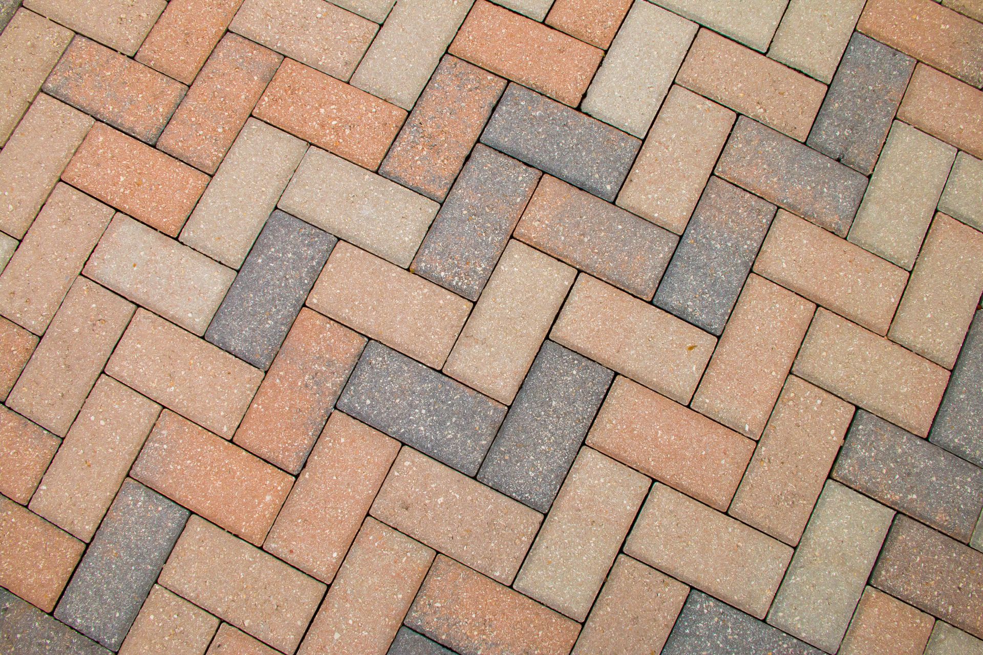 A herringbone pattern of rectangular paving stones in alternating shades of tan, brown, and grey.