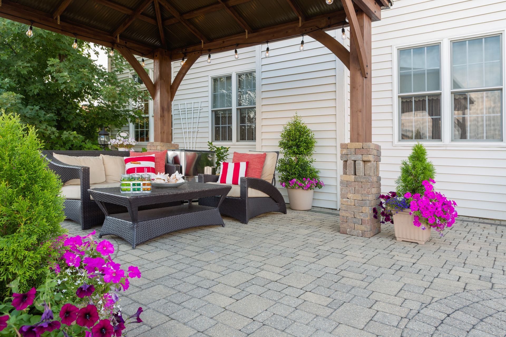 Patio seating under a wooden pergola with red-and-white cushions, stone pavers, and bright pink flowers.
