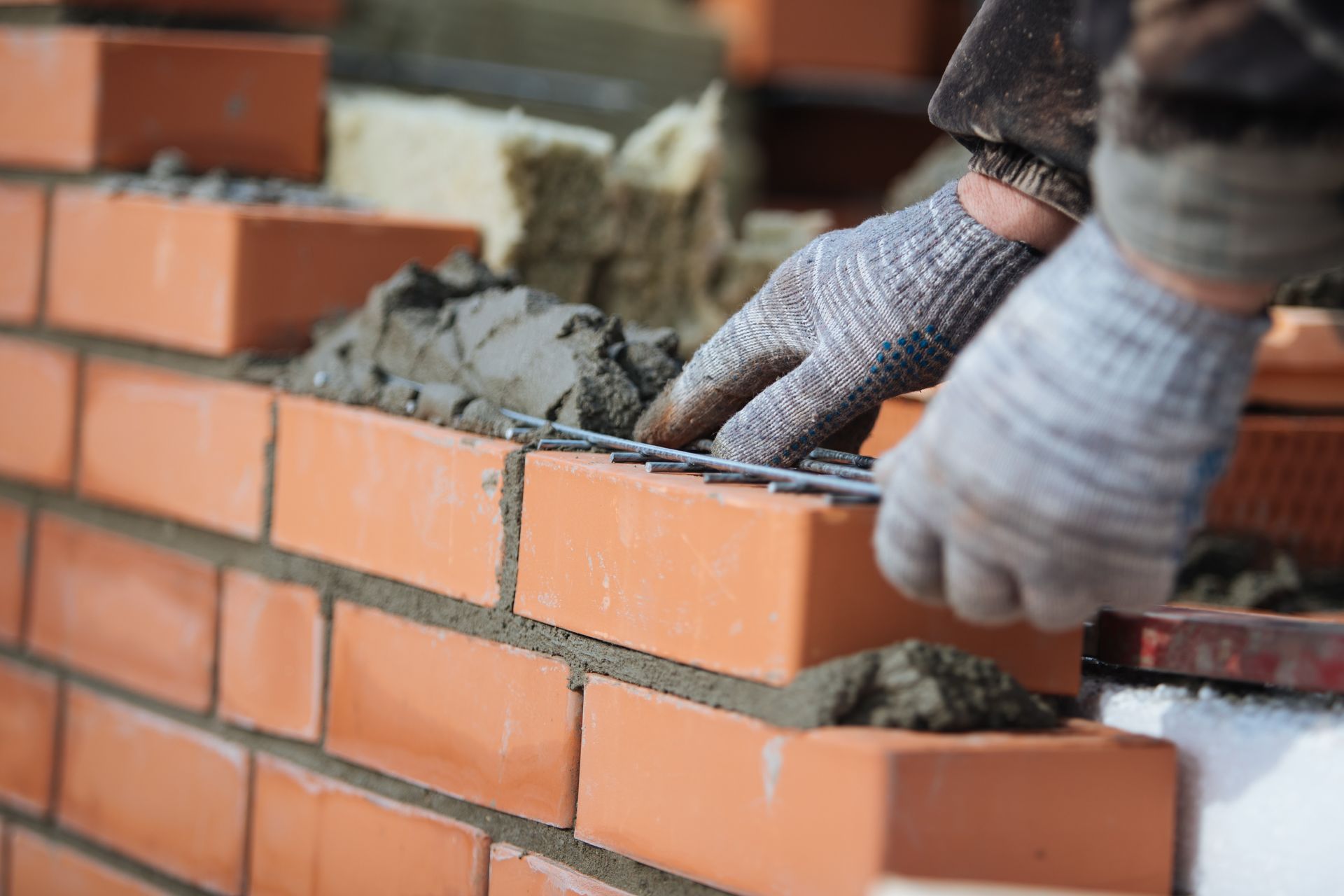 A worker in gray work gloves places metal reinforcement onto a layer of mortar between courses of red clay bricks.