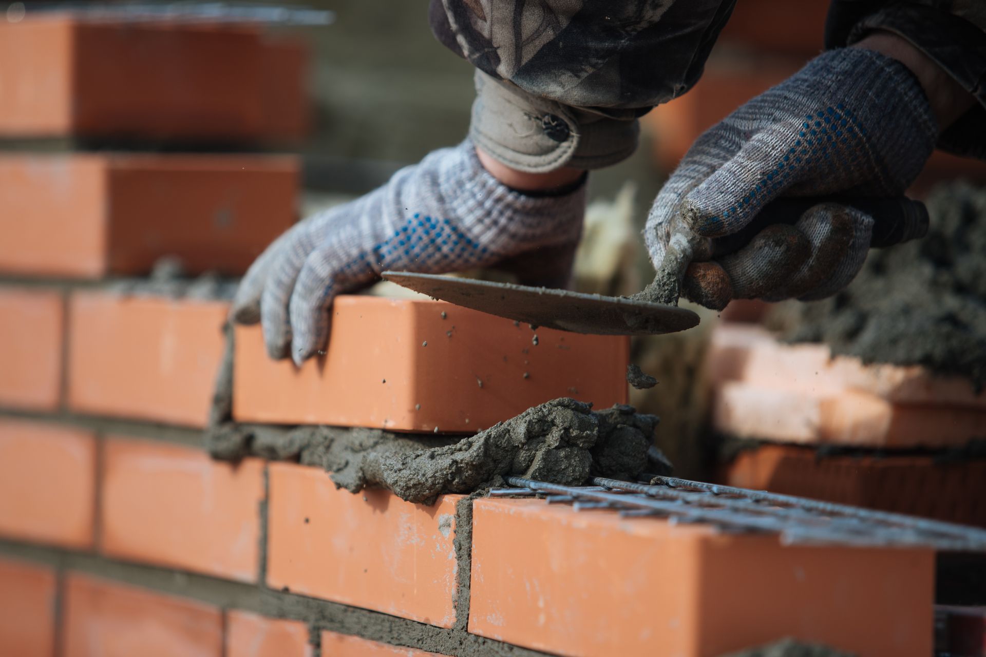 A gloved hand uses a trowel to apply mortar to a red brick while building a wall.