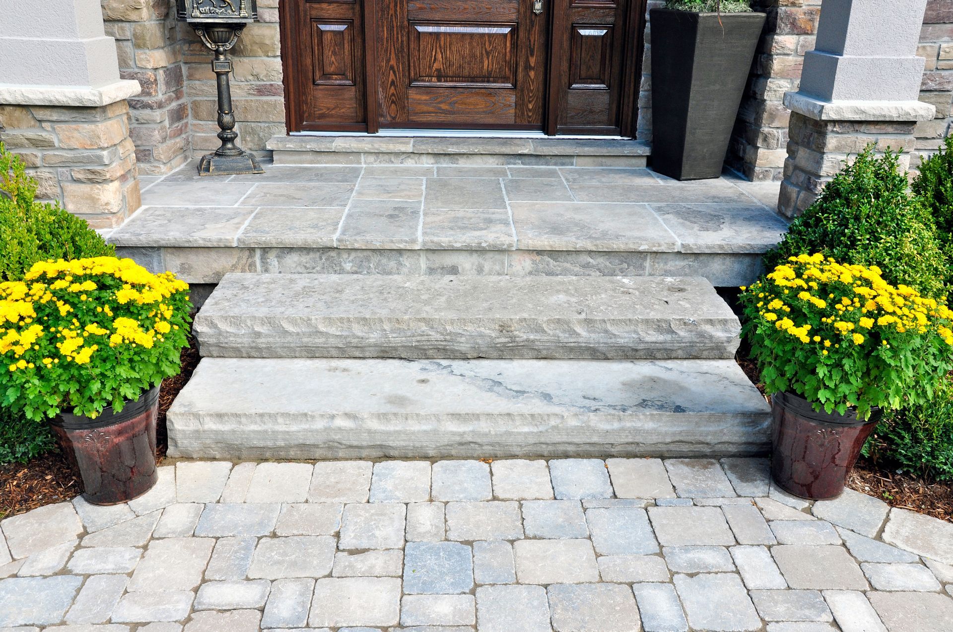 Stone steps leading to a front door, flanked by two potted yellow flower arrangements on a paved patio.