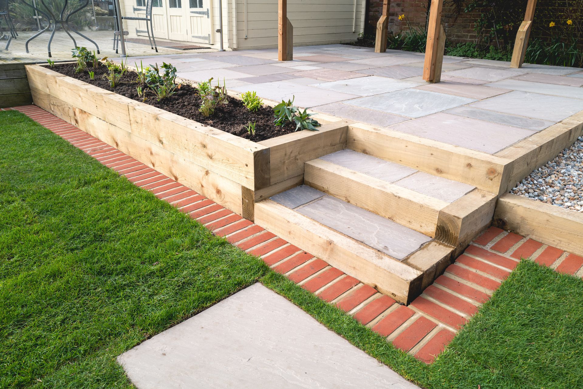 A raised timber garden bed with a planting area next to stone steps leading to a patio, bordered by red brick pavers.