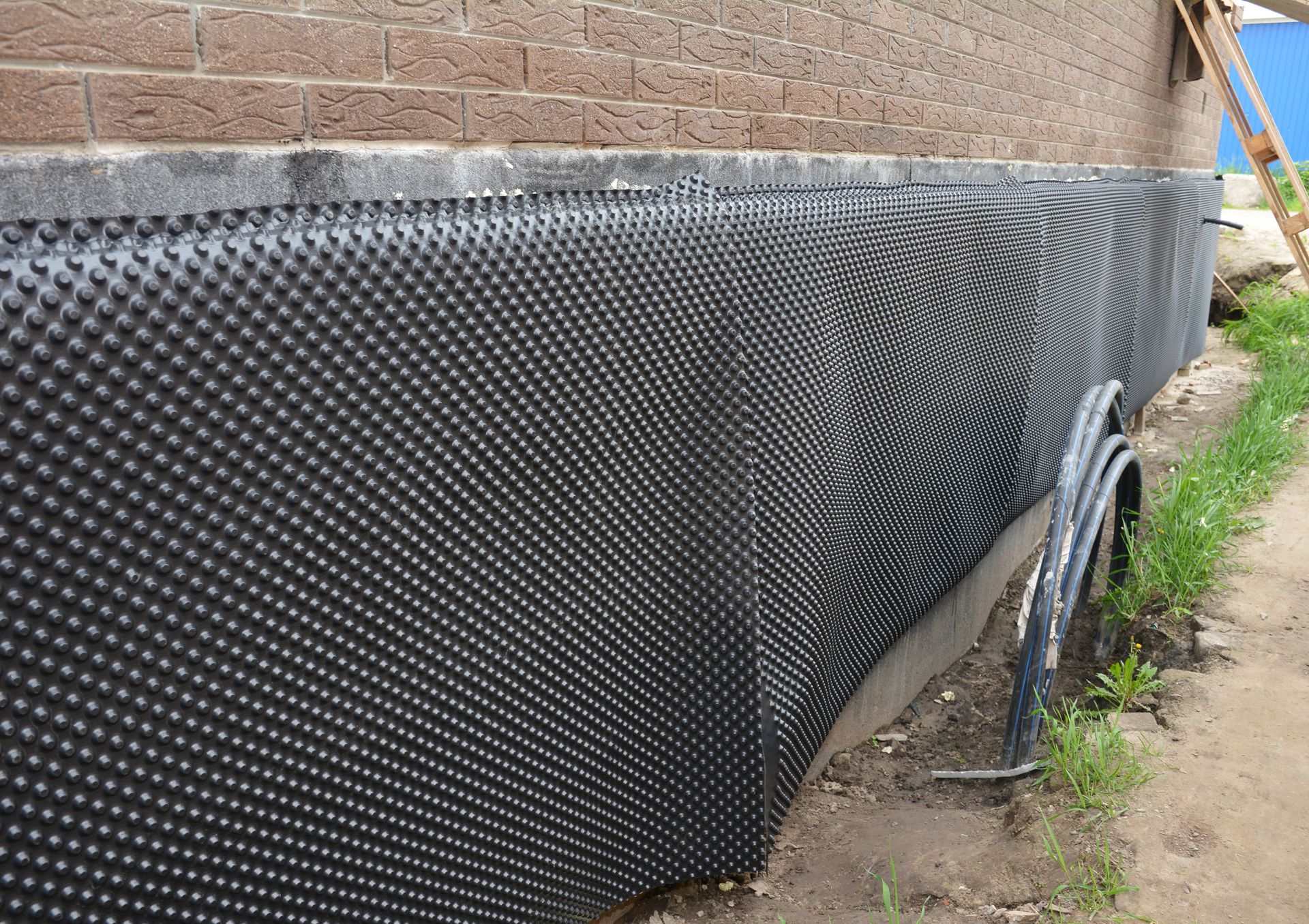 Black dimpled drainage membrane installed along a brick building foundation at ground level.