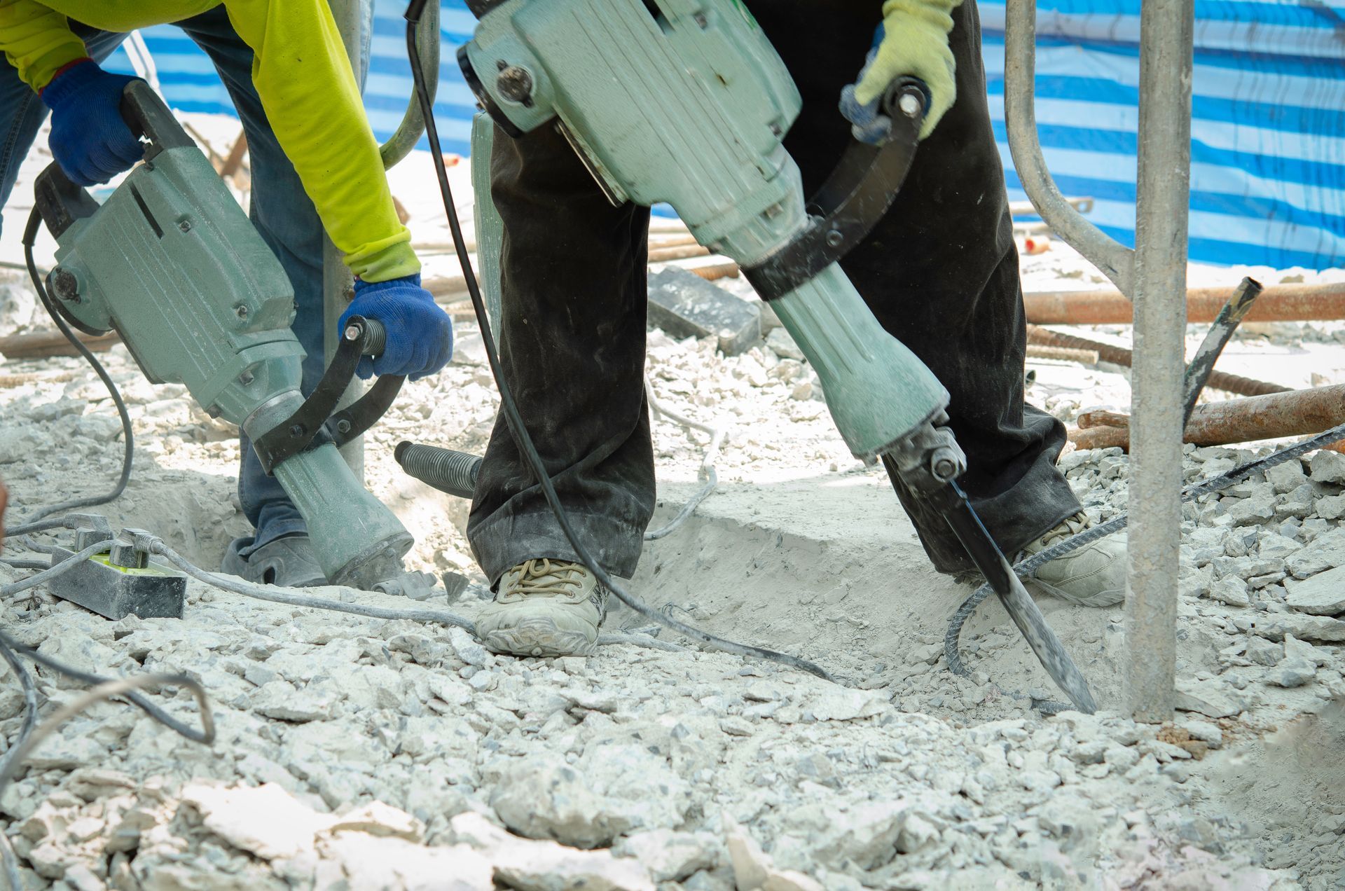 Two construction workers use heavy-duty jackhammers to break up concrete on a job site.