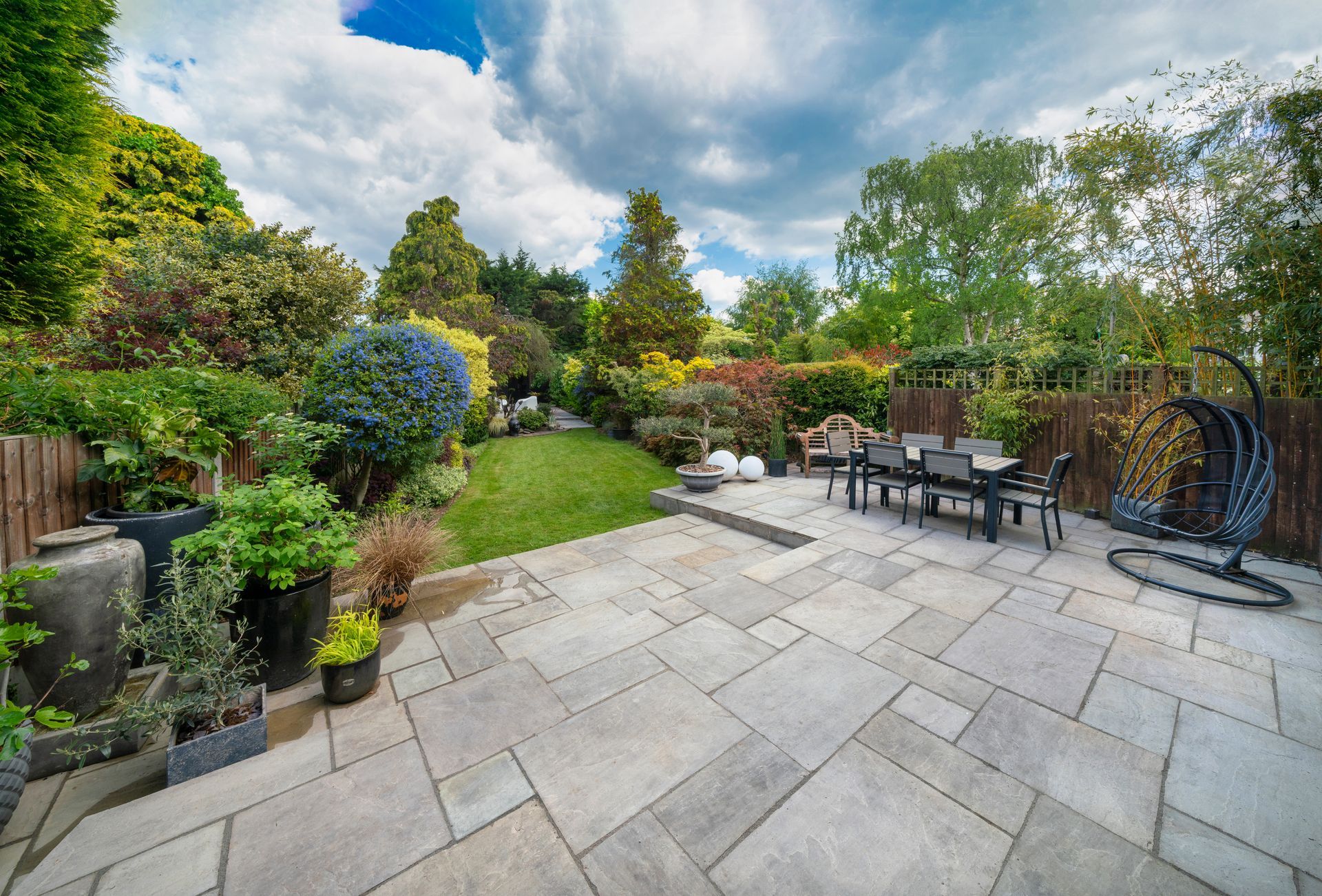 A sunny backyard patio features light-gray paving stones, outdoor dining furniture, a hanging chair, and lush greenery.