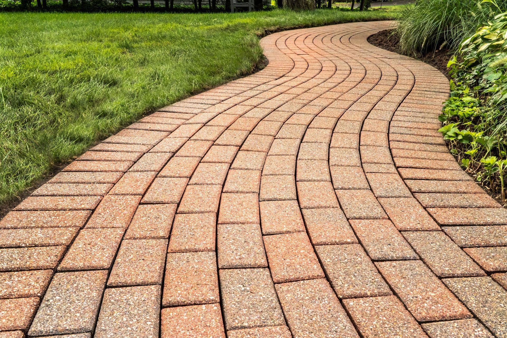 A winding path made of tan and reddish-brown rectangular paving stones, bordered by green grass and garden plants.