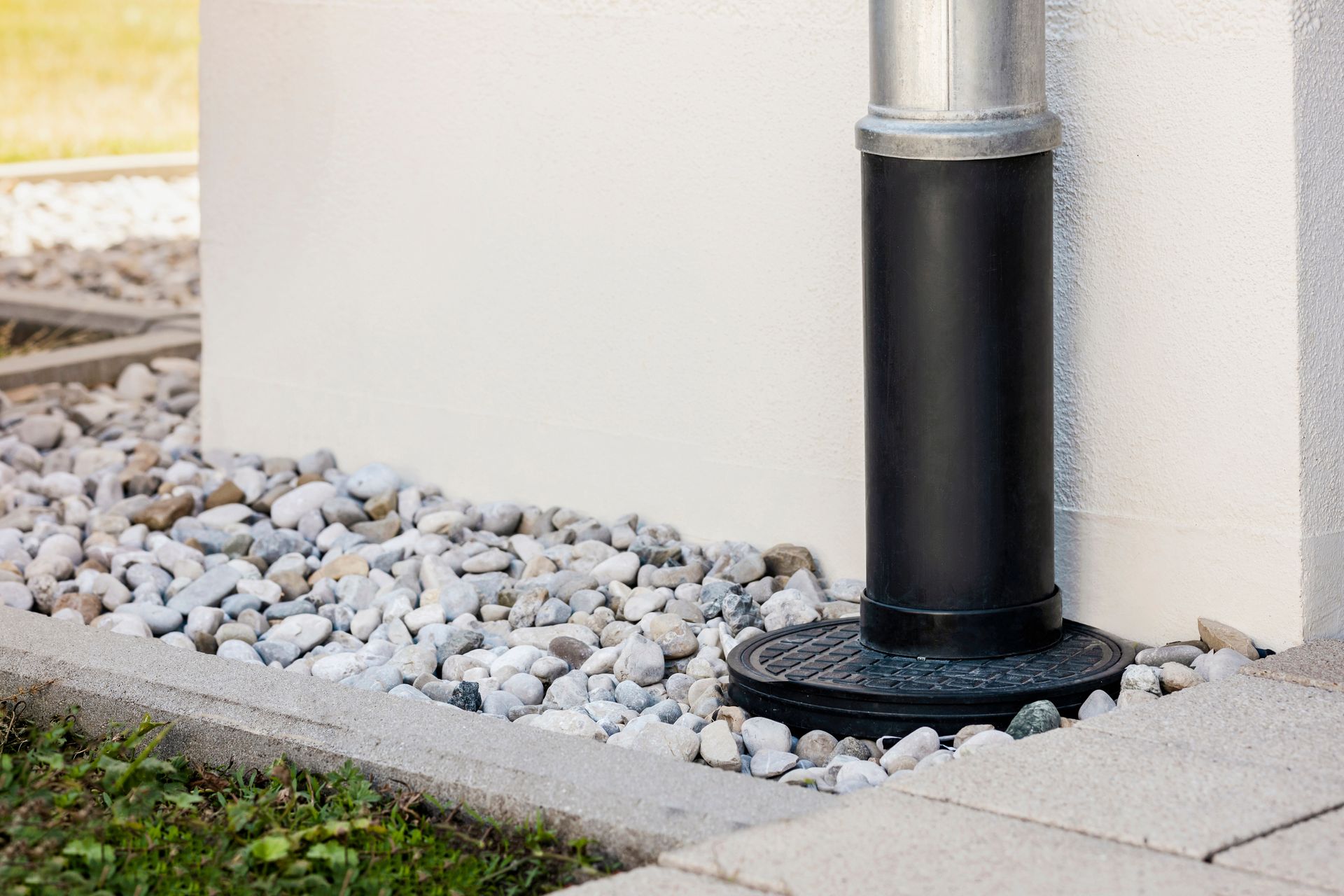 A metal and black pipe downspout connected to a ground-level drain, set against a light wall with a gravel border.