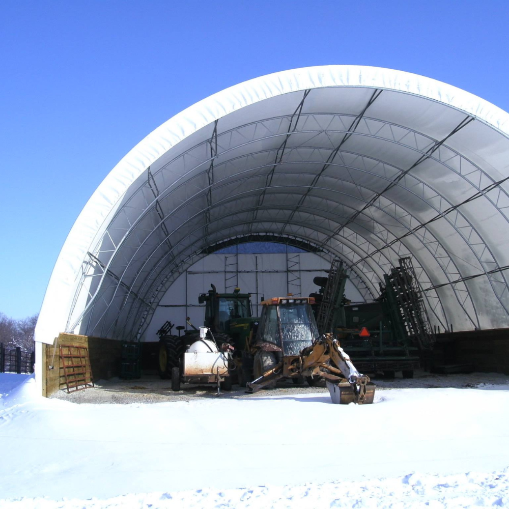 Large white arched storage structure protecting farm equipment in a snowy field.