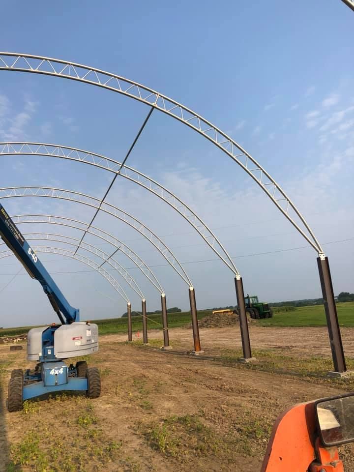 Arched metal framing for a structure under construction; blue lift on the left, open field in the background.