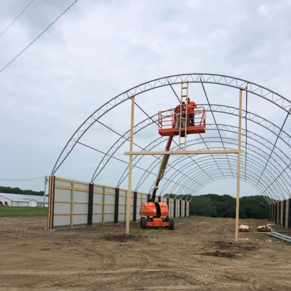 Construction of a greenhouse: worker in an orange lift attaching lumber to metal frame. Cloudy sky.
