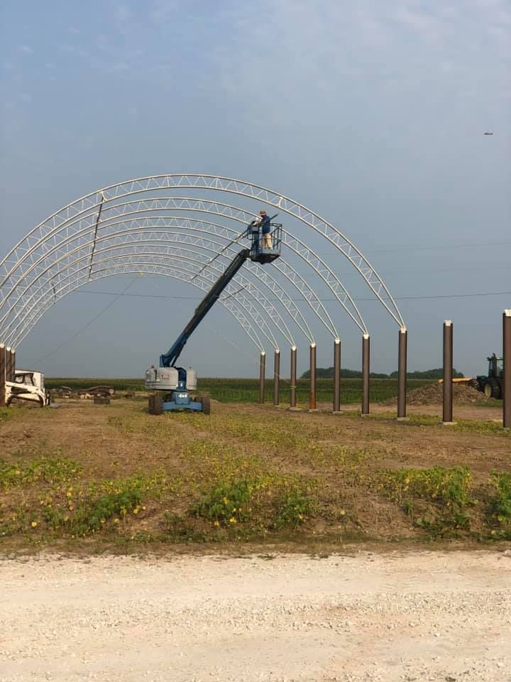 Construction worker on a lift assembling metal arches for a building frame on a field under a cloudy sky.