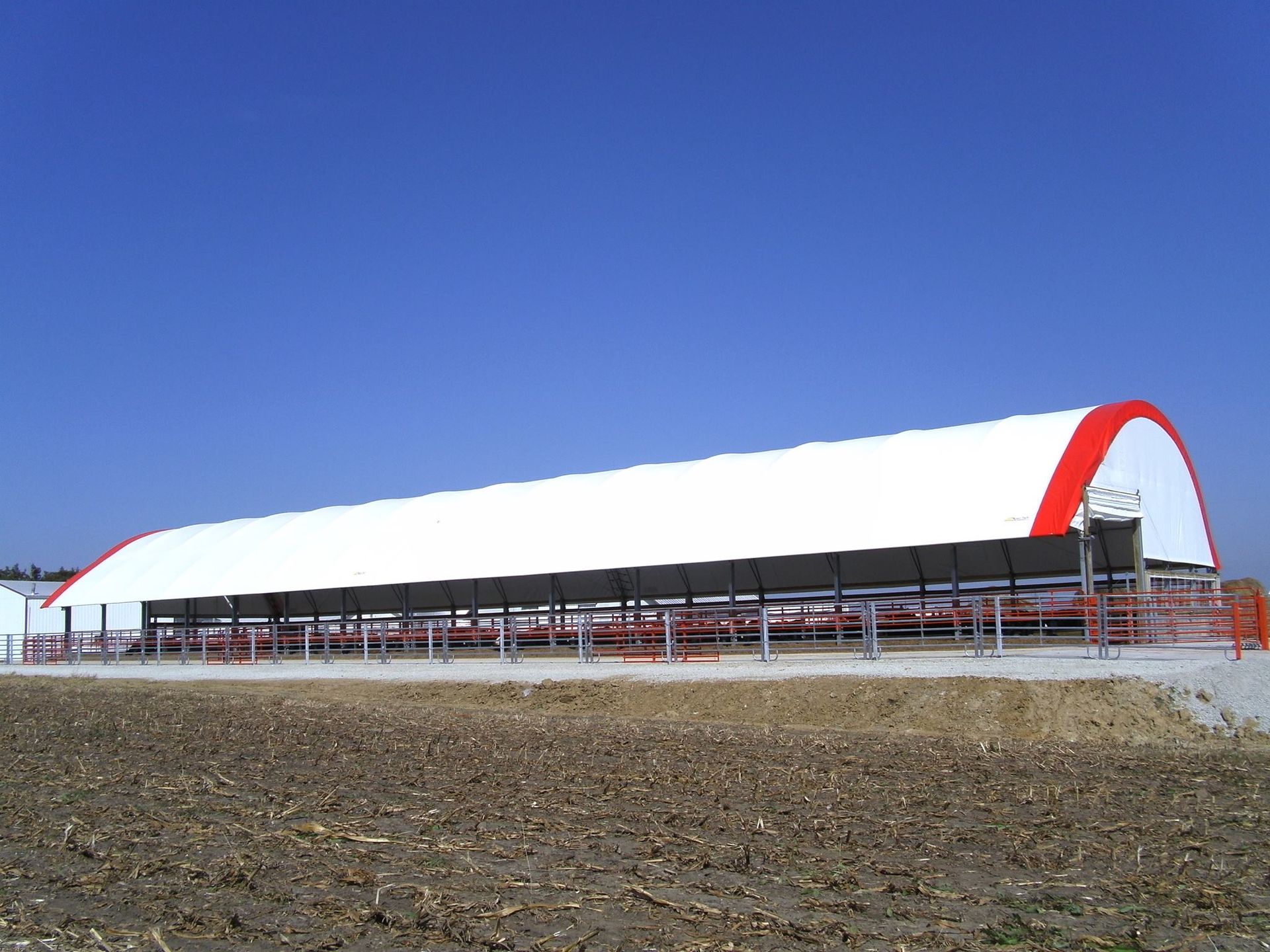 Long, white agricultural building with red ends, a black awning, and a blue sky.