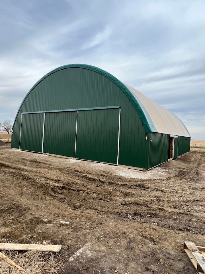 Green arched metal building with large sliding doors, on a dirt lot under a cloudy sky.
