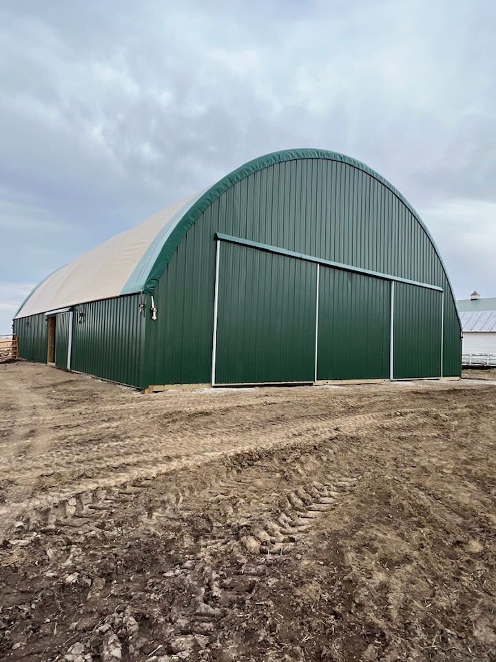 Green arched-roof metal building with large sliding doors on a dirt lot under a cloudy sky.