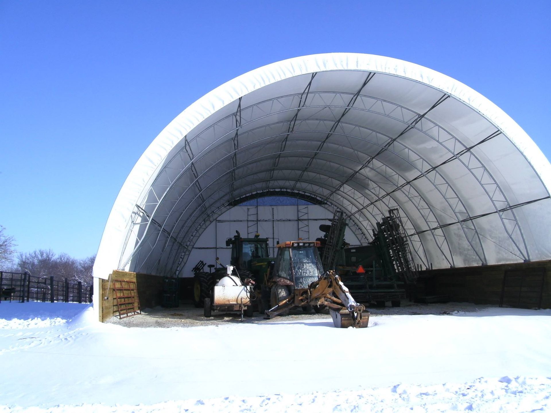 Large white arched shelter with machinery inside, set against a snow-covered landscape under a clear blue sky.