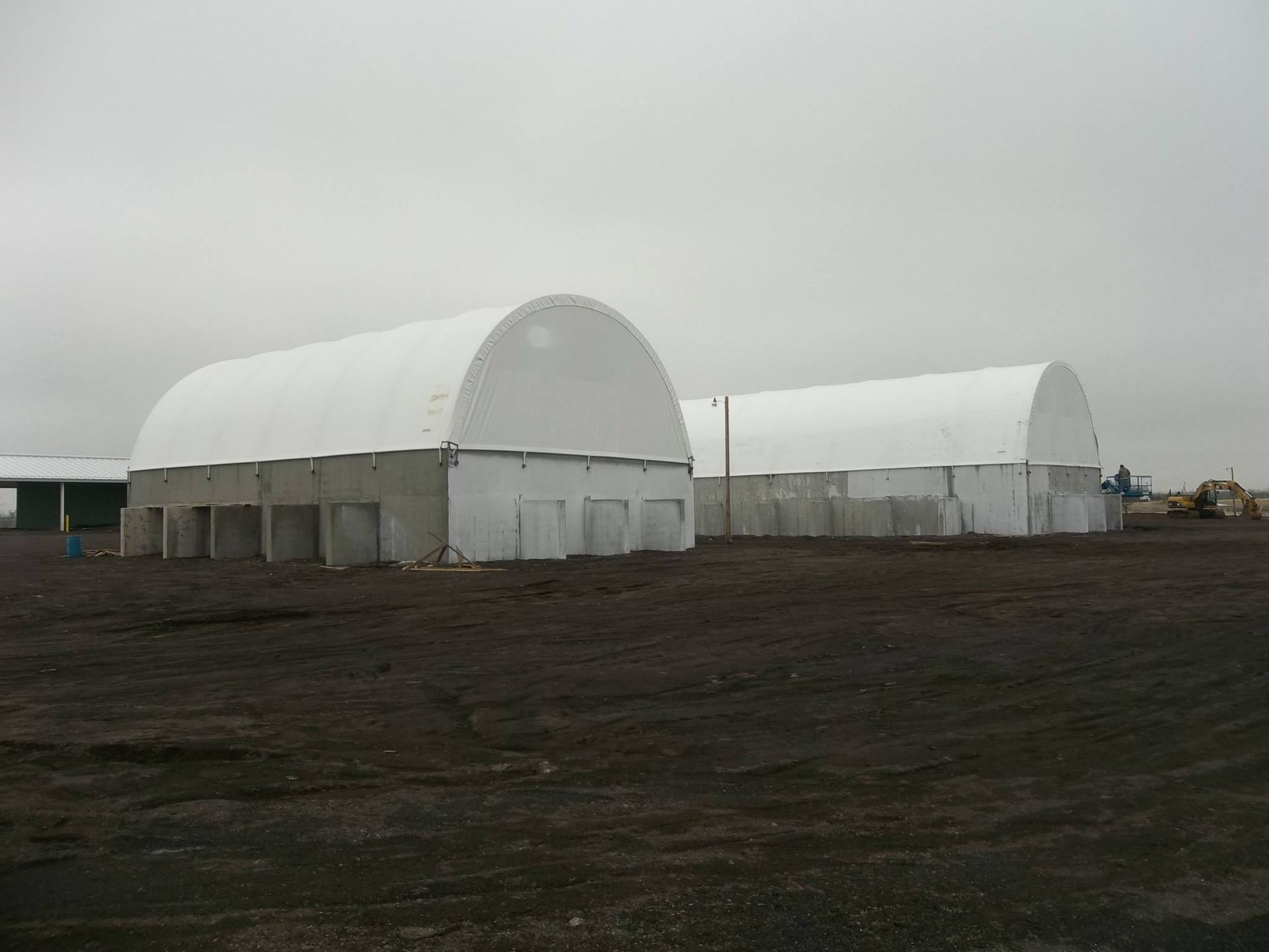Two white arched roof buildings with concrete bases on a dark field under a cloudy sky.