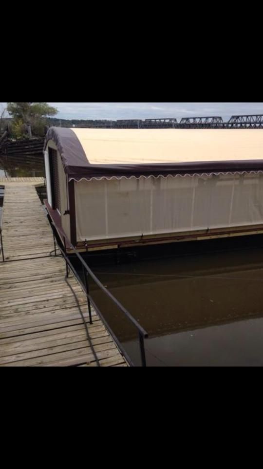 Houseboat docked next to a wooden pier on the water with tan roof and white and brown sides.